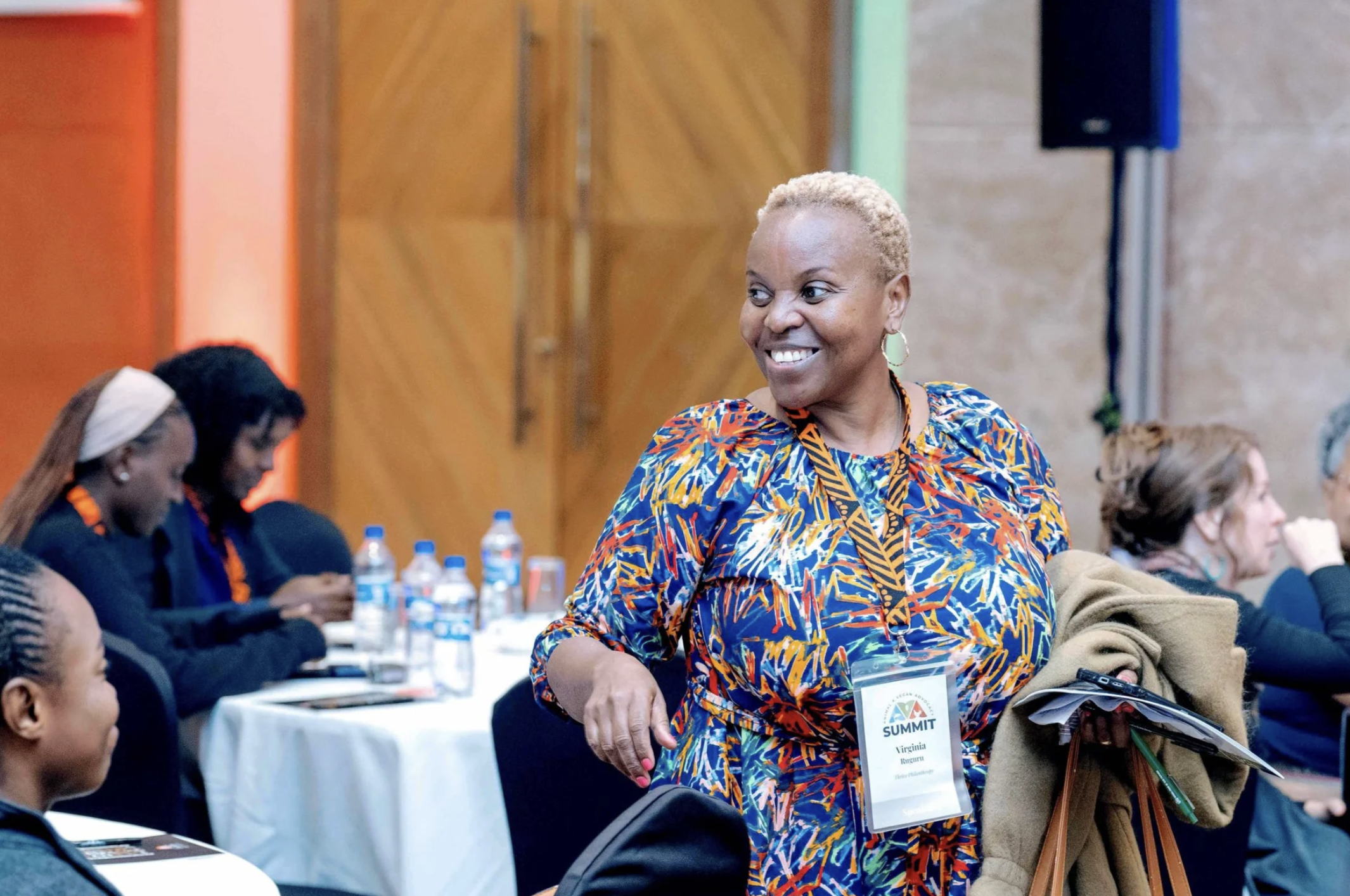 A smiling woman with short blonde hair wearing a colorful patterned dress and a lanyard, attending a conference or seminar, holding a coat and some papers.