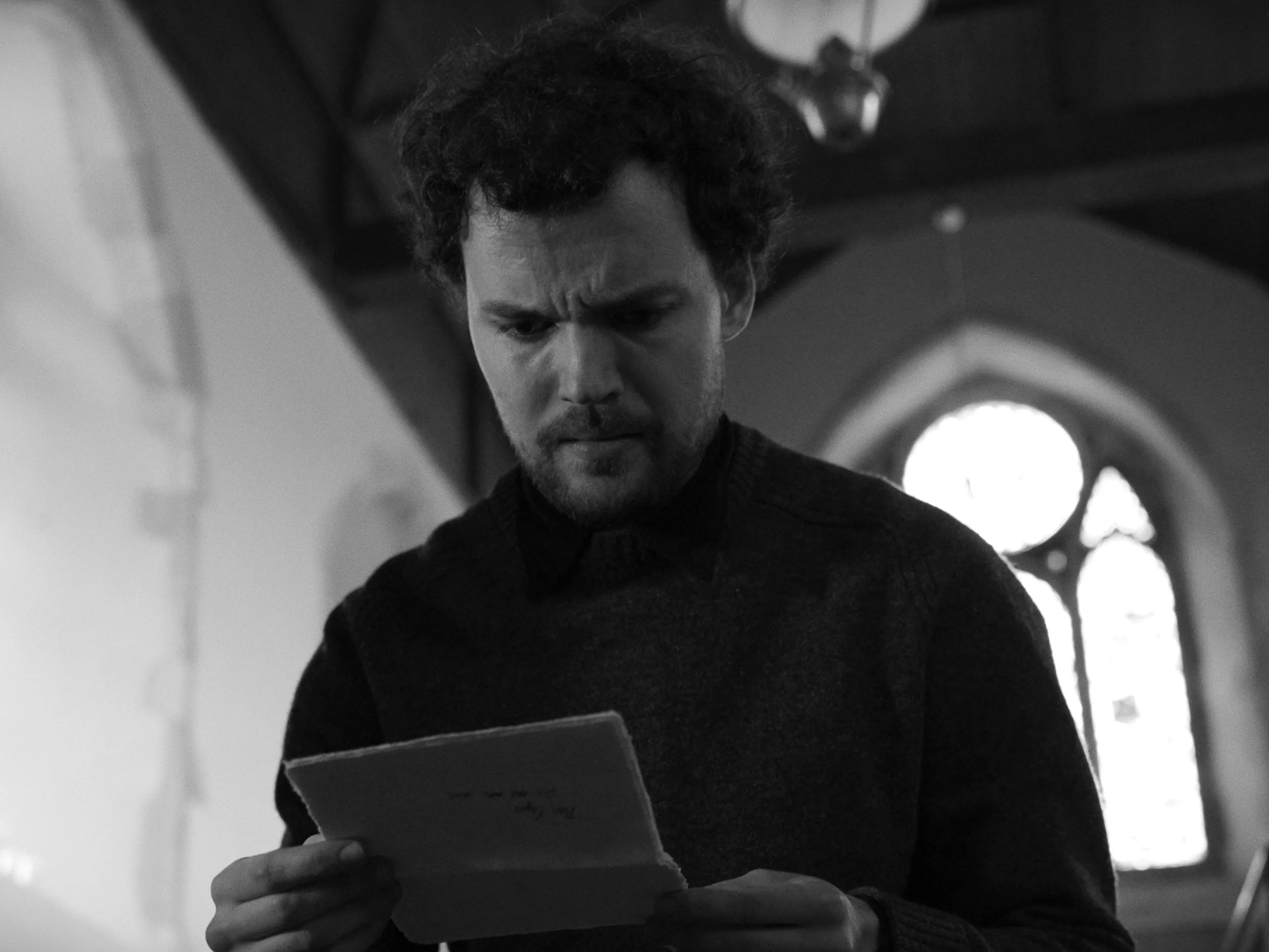 A man with curly hair and a beard looking down at a tablet or phone he is holding in his hand. The scene appears to be indoors, possibly in a church or cathedral, with round windows and ornate ceiling details.
