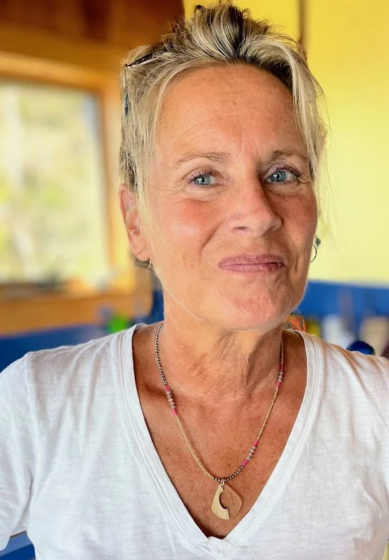 A woman with short blonde hair, wearing a white shirt and a necklace, making a playful facial expression in an indoor setting with yellow and blue walls.