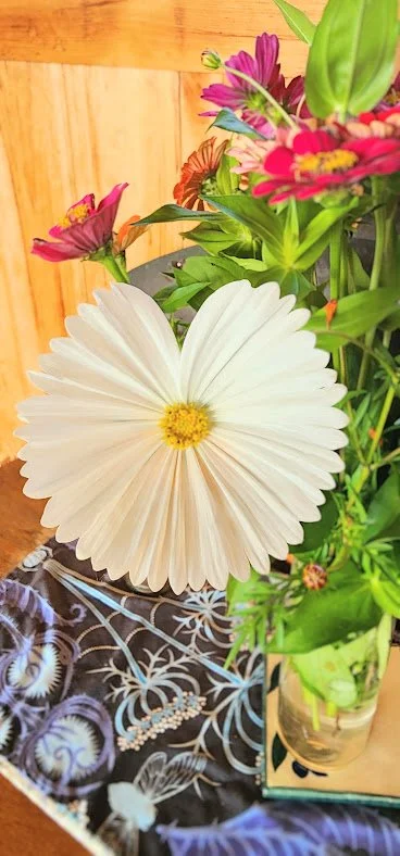 A white pleated paper fan in the shape of a flower, surrounded by pink and purple flowers, and green leaves in a glass vase, on a table with a patterned cloth.