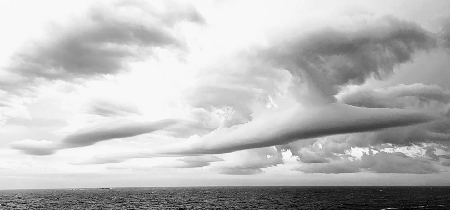 Black and white photo of overcast sky with large, dark clouds above a calm ocean horizon.
