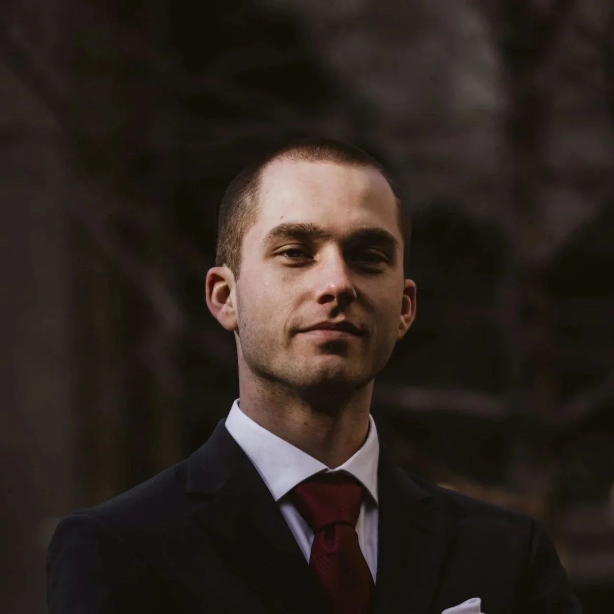 A young man with short dark hair wearing a black suit, white shirt, and red tie, standing outdoors with a blurred natural background.