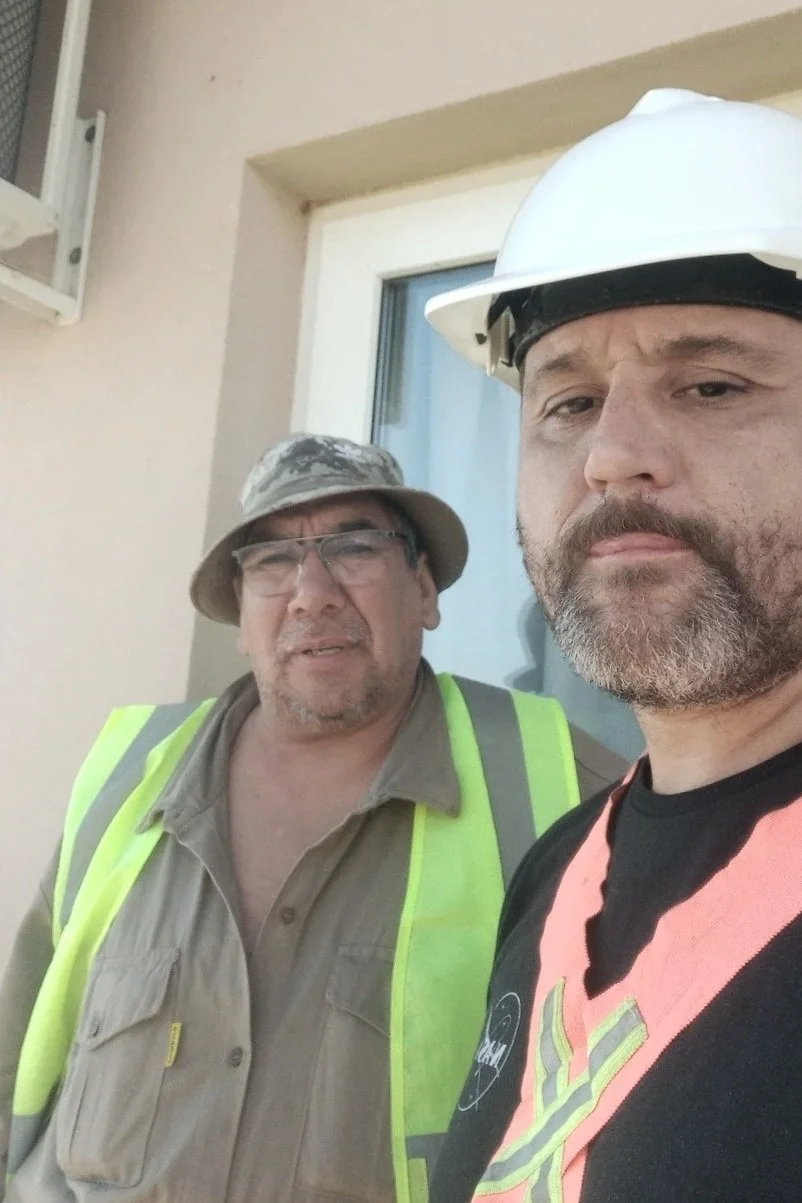 Dos hombres en equipo de construcción, uno con casco blanco y el otro con sombrero y gafas, posando juntos