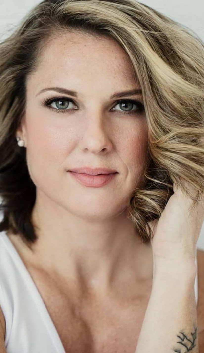 Close-up of a woman with wavy blonde hair, blue eyes, wearing pearl earrings and a white top.