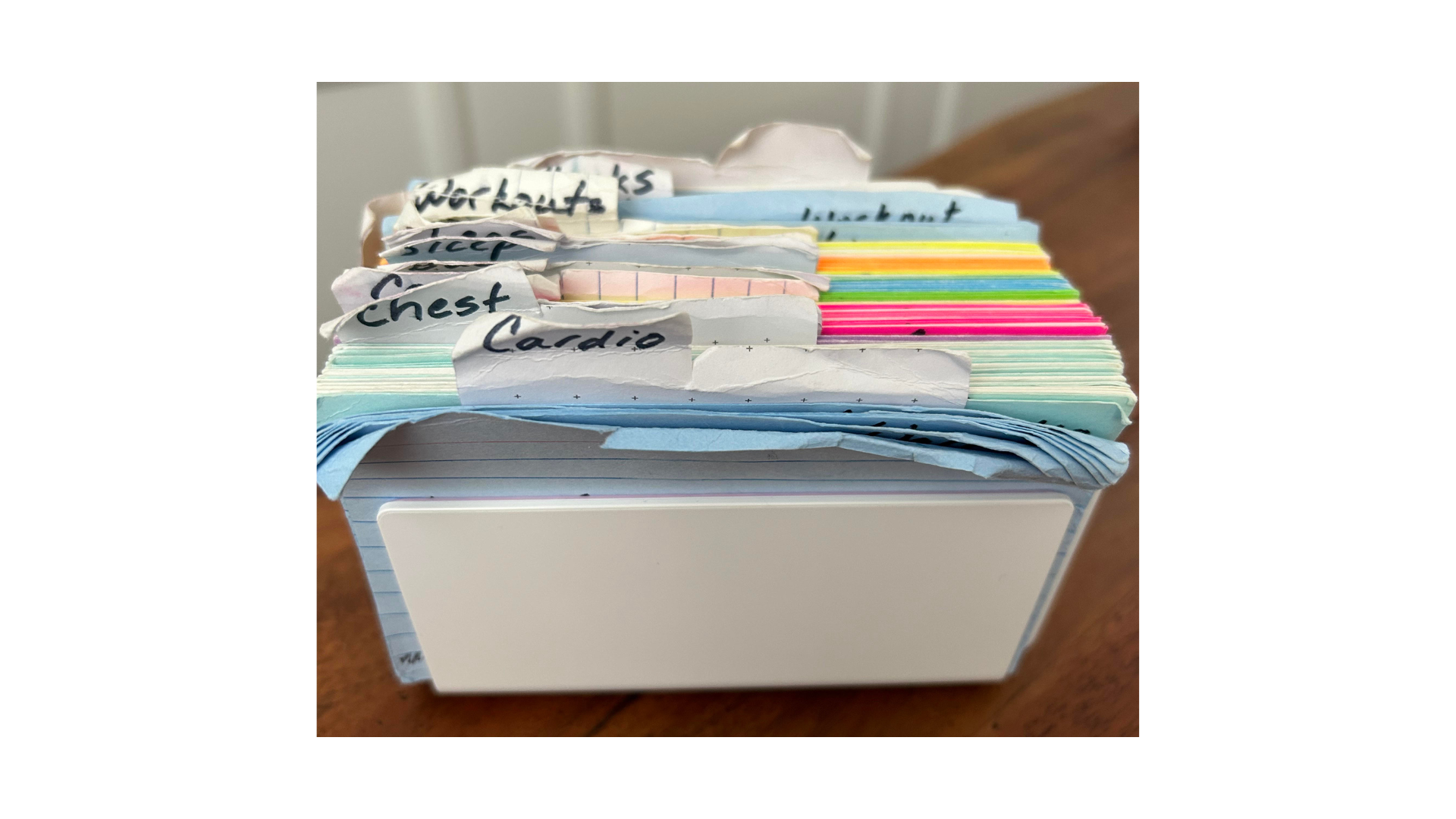 A white box filled with crumpled and organized index cards labeled with workout names, including 'Chest,' 'Cardio,' and 'Workouts,' resting on a wooden surface.