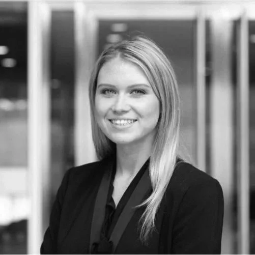 Black and white photo of a woman with straight blonde hair wearing a black blazer, smiling at the camera, in an indoor setting.