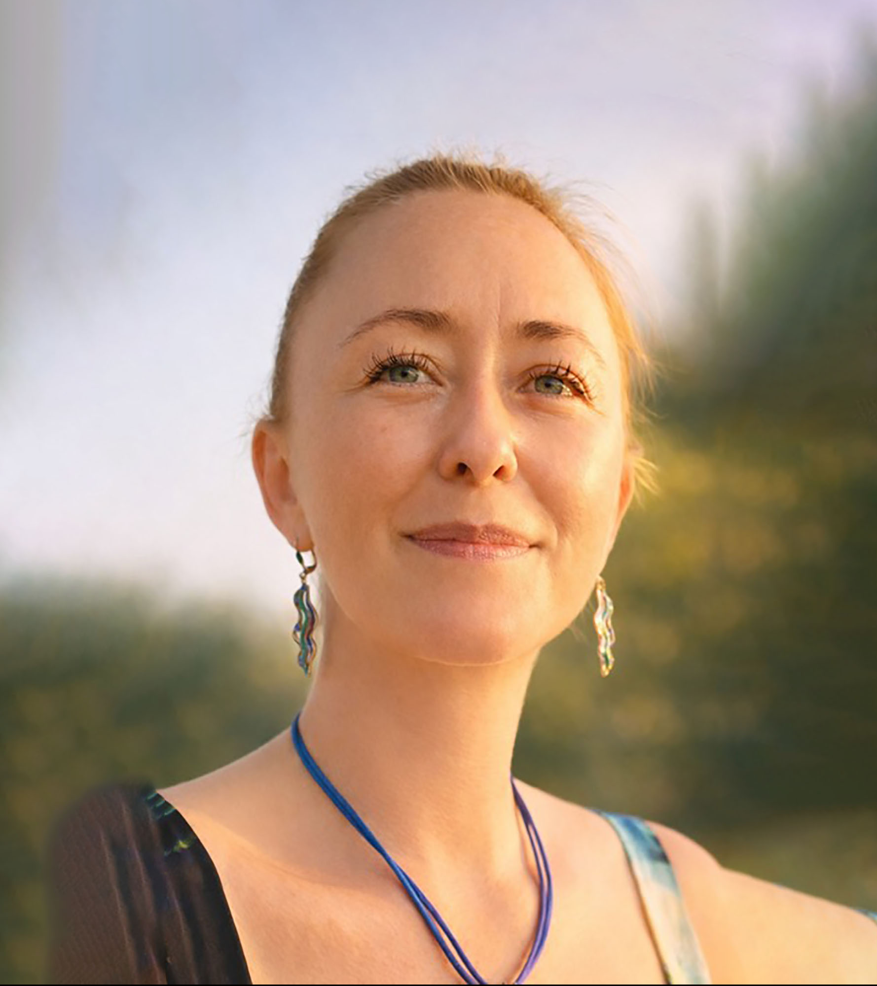 Nahaufnahme einer lächelnden Frau mit blonden Haaren, blauen Augen und Ohrringen, die im Freien bei Sonnenuntergang steht.