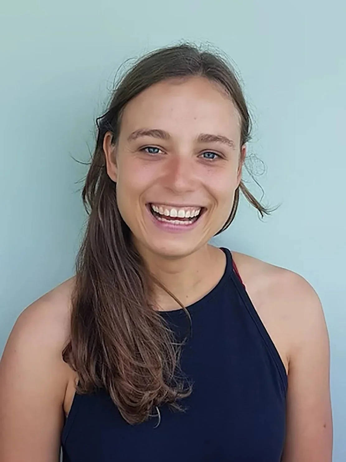 Lächelnde junge Frau mit braunen Haaren und blauen Augen, trägt ein dunkles Sporttop, steht vor einer hellen Wand.
