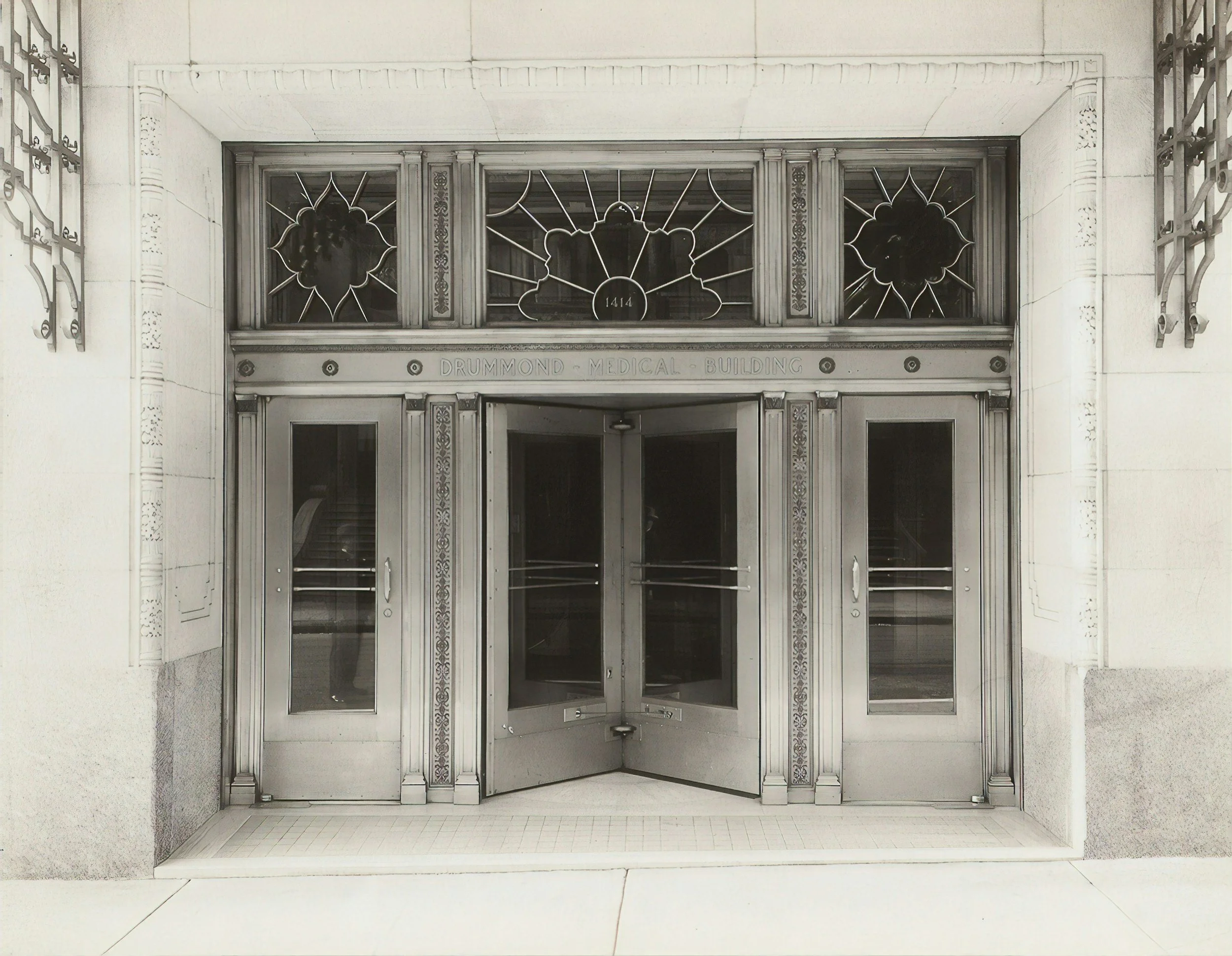 Black and white photo of the entrance to Drummond Medical Building, featuring a glass door with an ornate metal frame and decorative stained glass windows above.