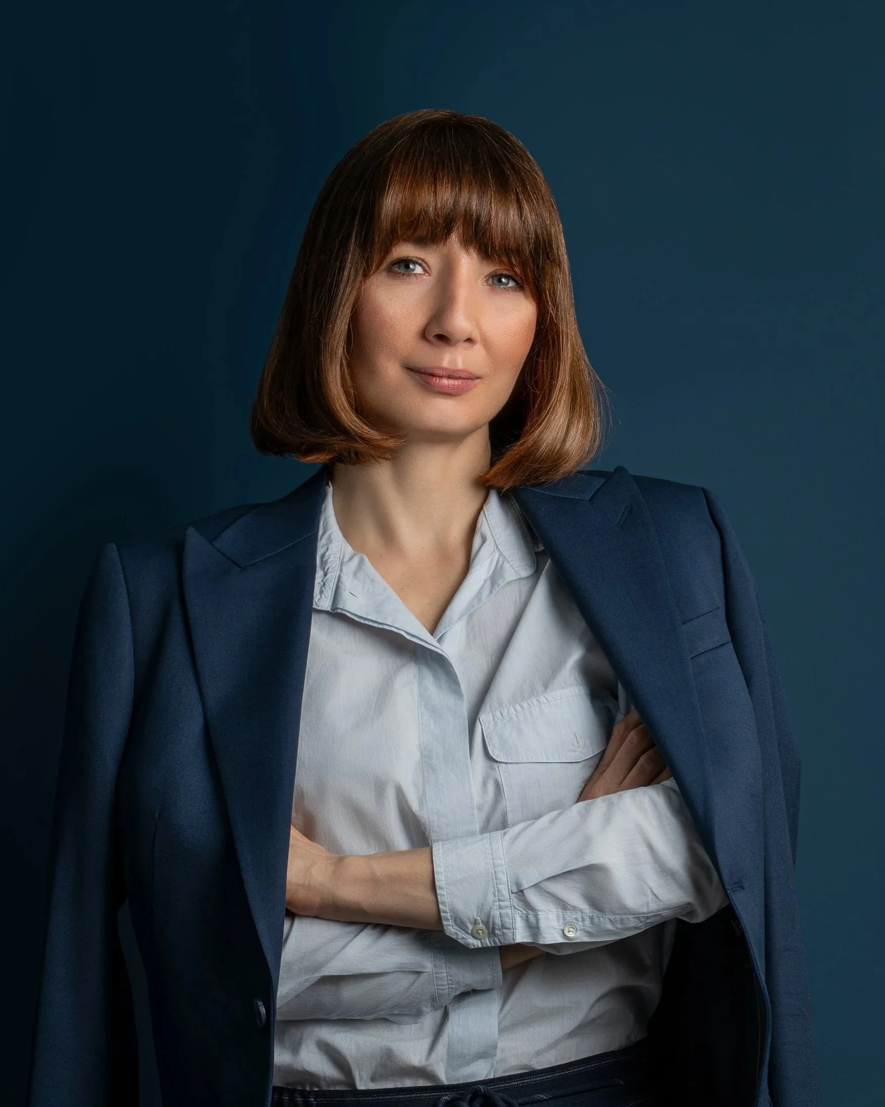 Portrait of a woman with shoulder-length brown hair, wearing a white shirt and a blue blazer, standing against a dark blue background.
