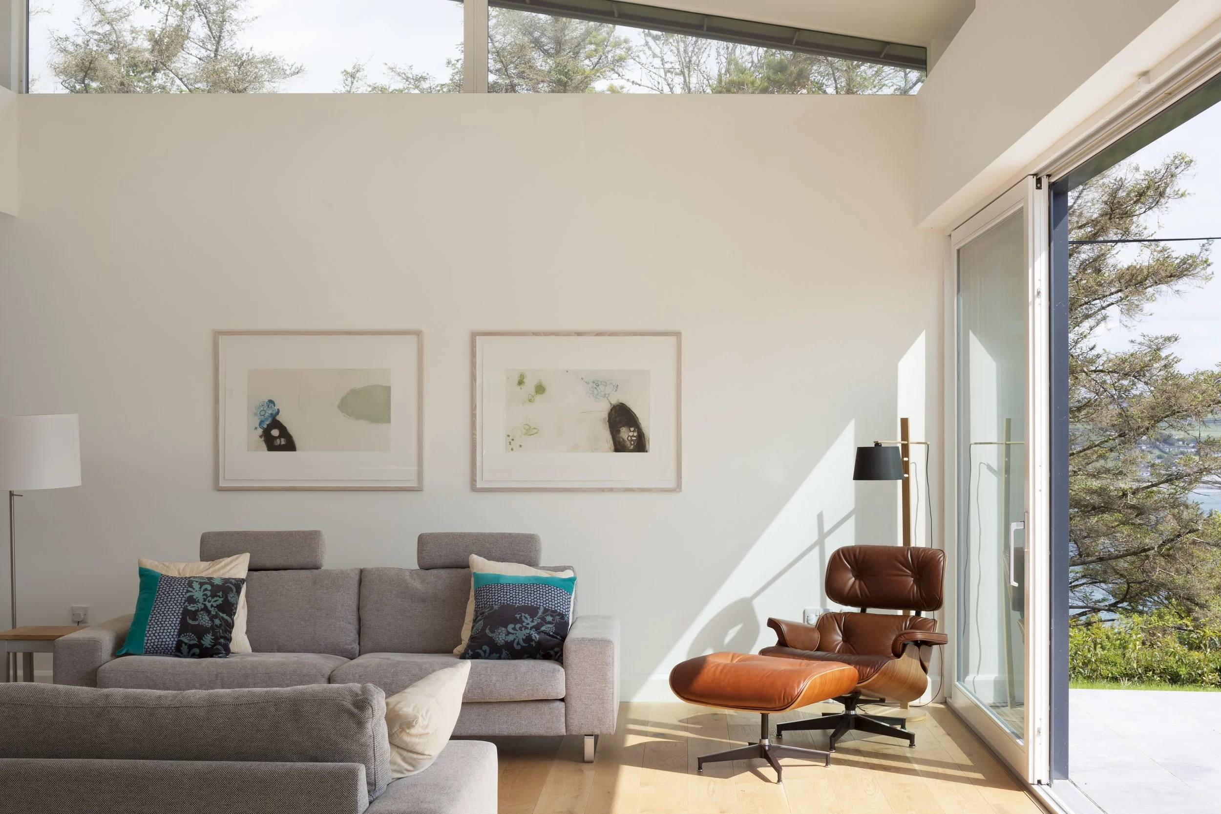 Interior view of a modern living room with a large window, a gray sofa with colorful pillows, a brown leather lounge chair, and wall art featuring abstract portraits, overlooking a garden with trees.