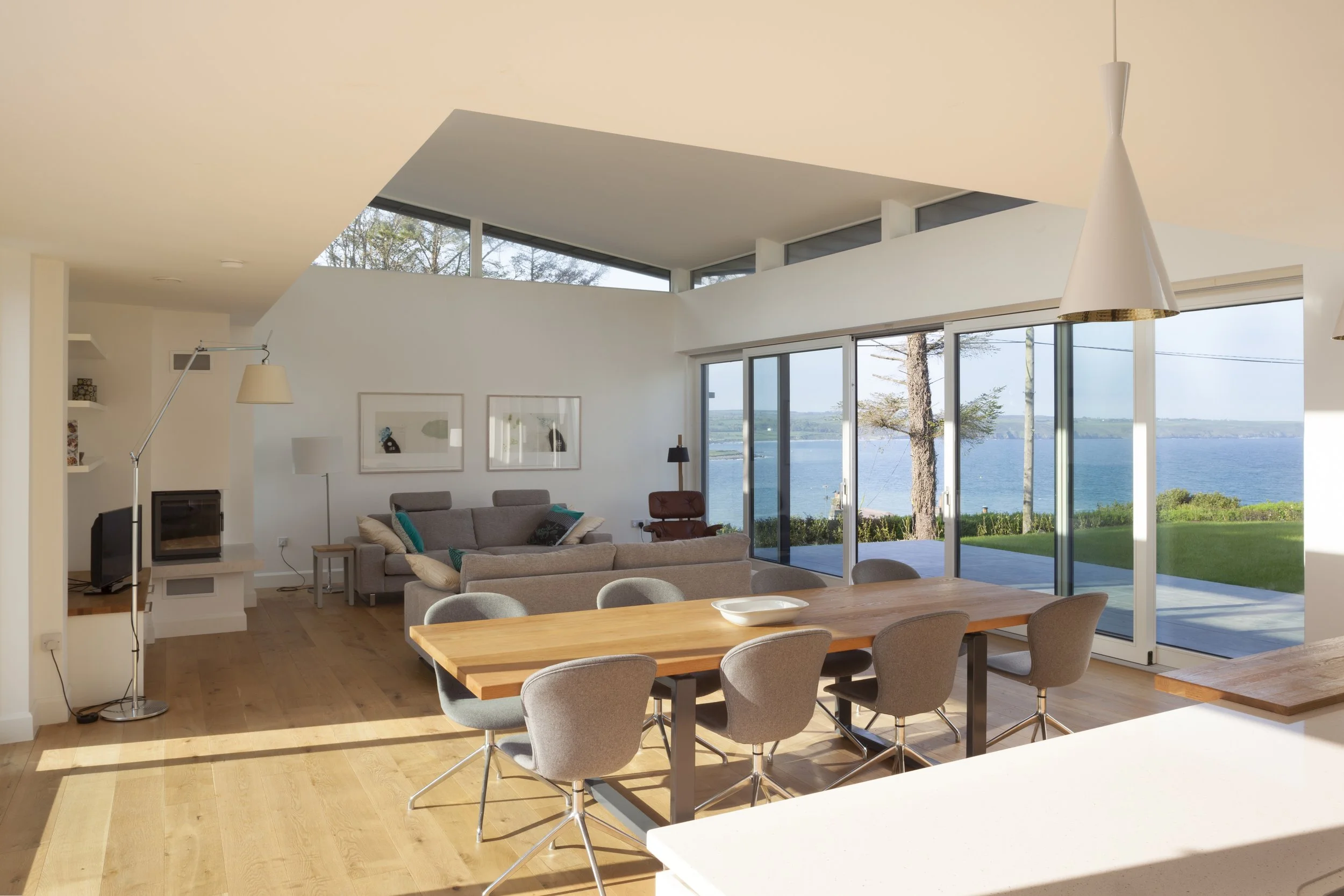 Modern living room with large windows overlooking water, beige sofas, a wooden dining table with gray chairs, and indoor lamps.