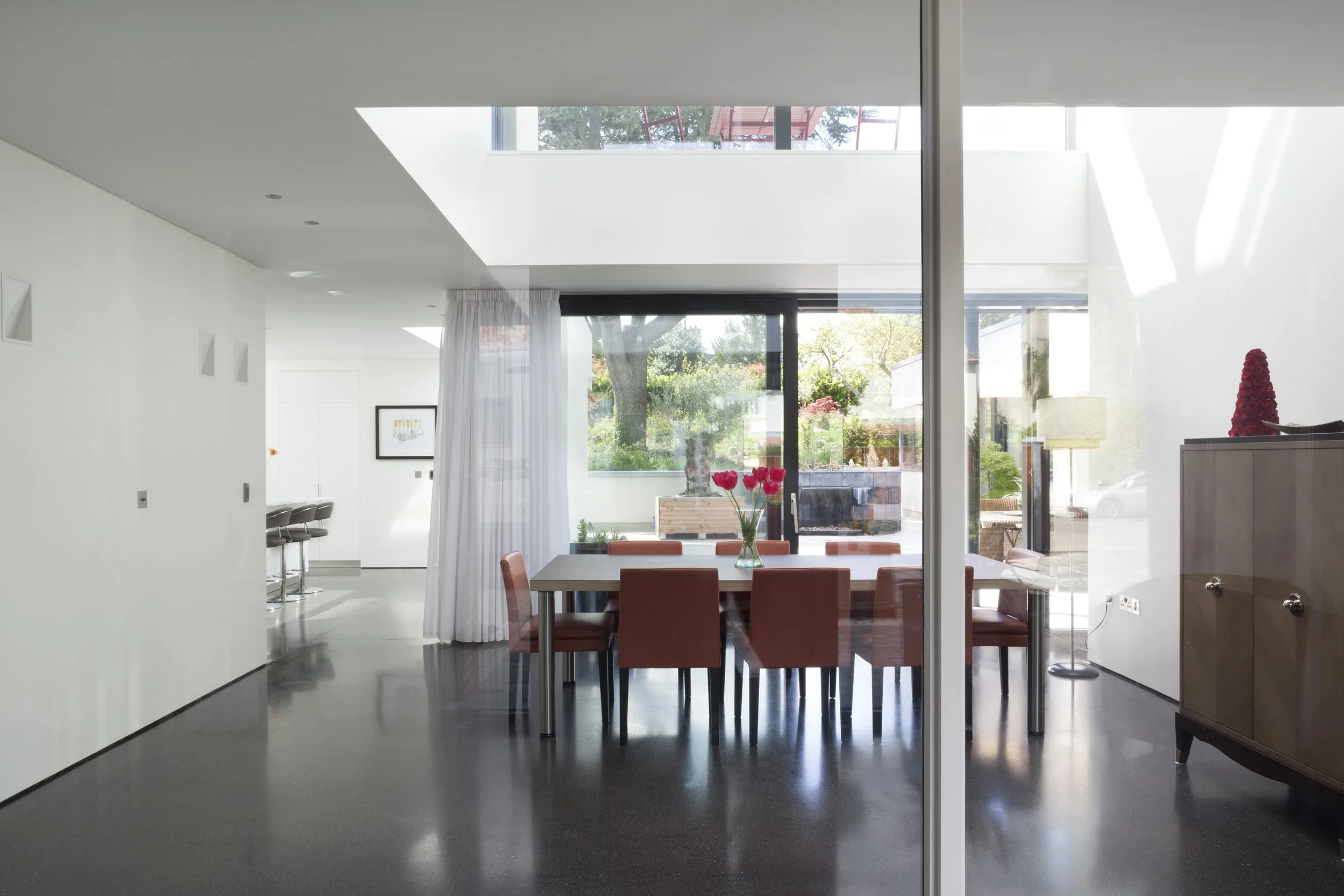 Modern dining area with a wooden table, terracotta chairs, and a vase with pink tulips. Large windows reveal a garden with trees and outdoor furniture. Bright, minimalist interior with white walls and ceiling.