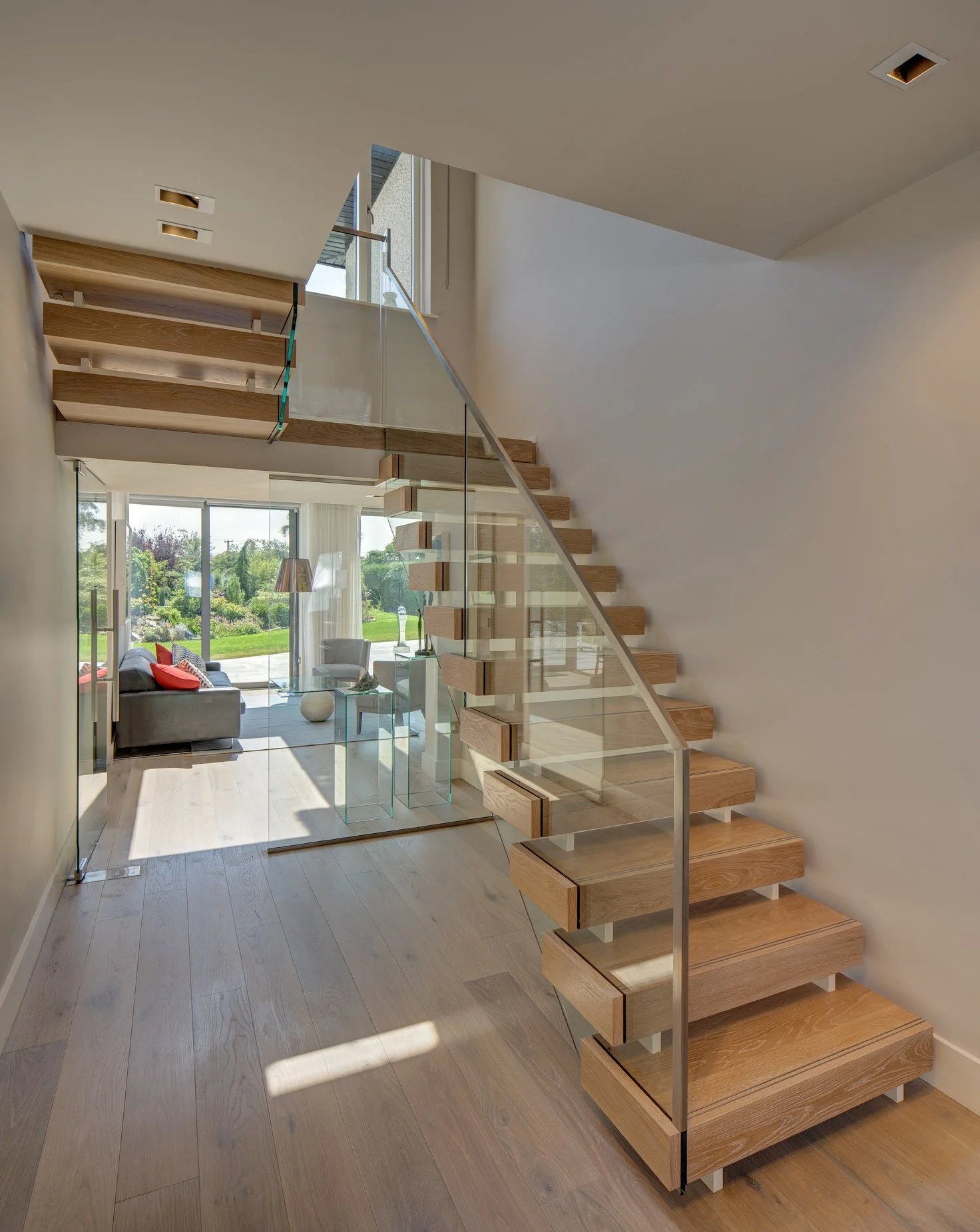 Modern interior with floating wooden stairs, glass railing, and a view of a living room with large sliding glass door leading to a landscaped backyard.