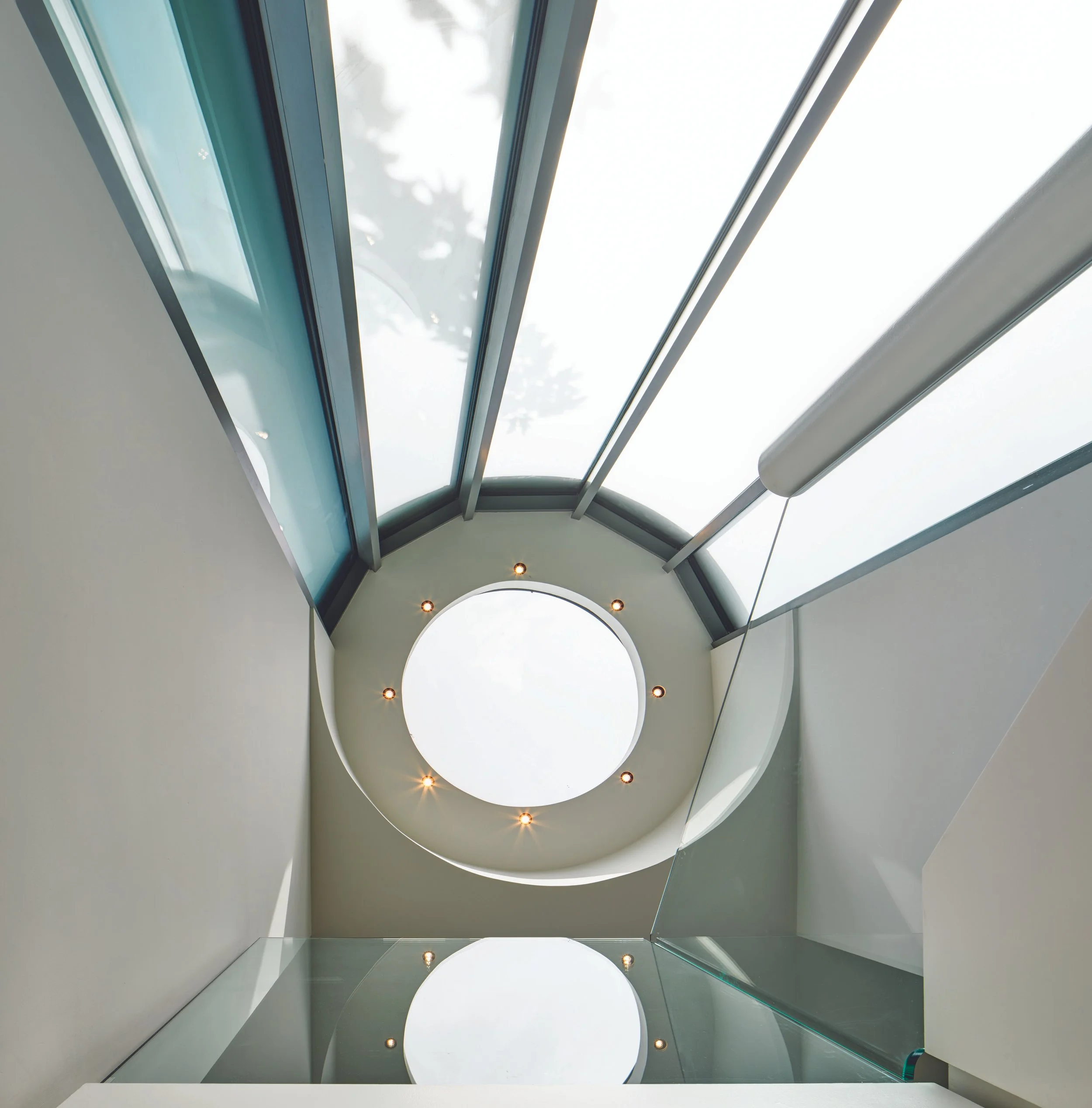 Looking up at a circular skylight in a modern interior ceiling. The ceiling has curved white walls with recessed lighting around the circular opening, allowing natural light to illuminate the space.