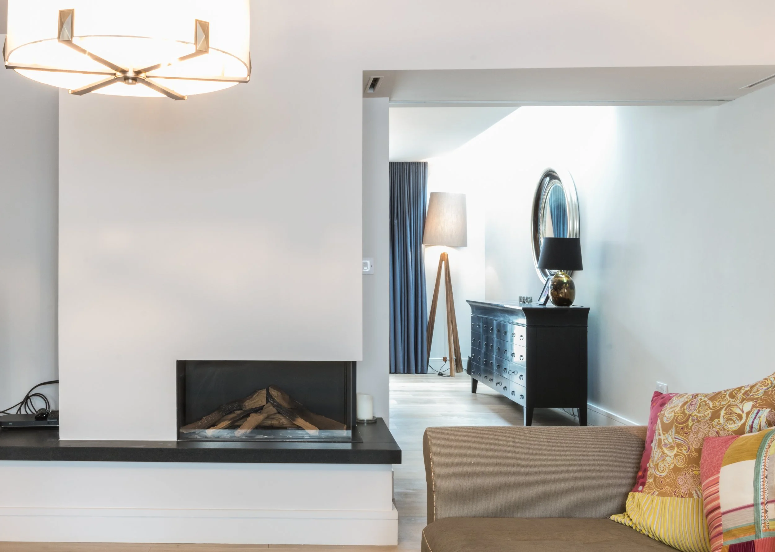 Modern living room with a fireplace, a sofa with colorful pillows, a console table with a lamp and decorative items, and a partial view of a mirror and a curtain.