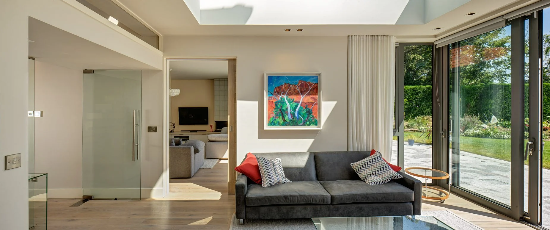 Modern living room with a dark gray sofa topped with red and patterned pillows, large sliding glass doors leading to a garden, white walls with colorful artwork, and a view into another room with a TV.