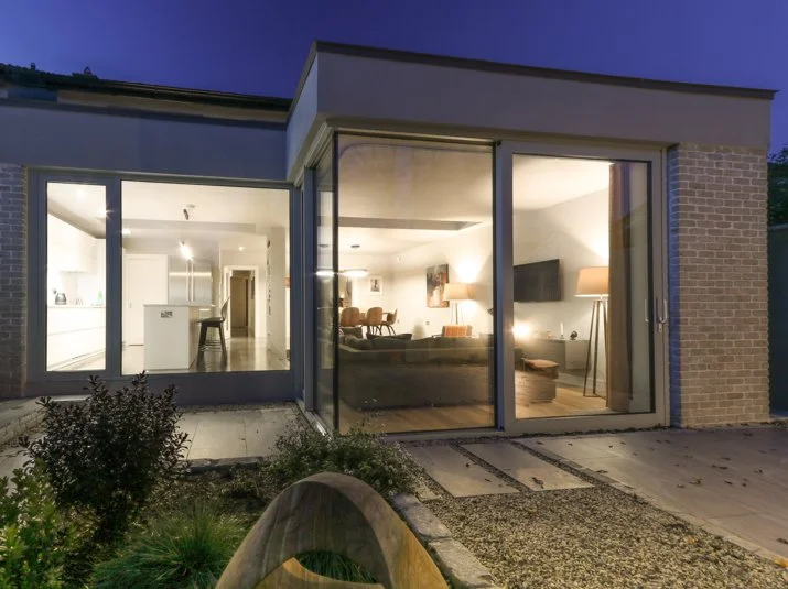 View of a modern house interior through large glass sliding doors, showing a living room with a couch, lamps, and a television, with a kitchen and dining area in the background.