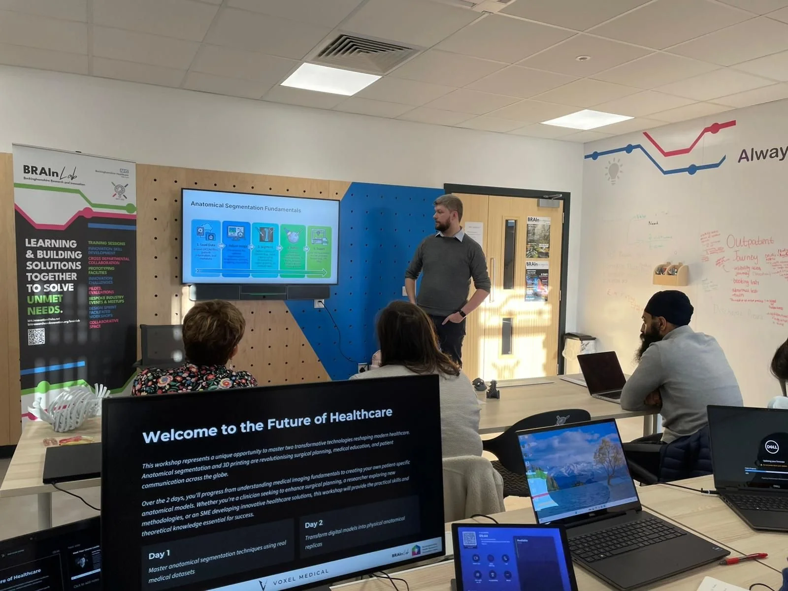 A man giving a presentation in a classroom with several attendees seated at tables. A large digital screen displays the presentation content, and there are multiple laptops open on the tables. The classroom has banners and posters related to healthcare and innovation.
