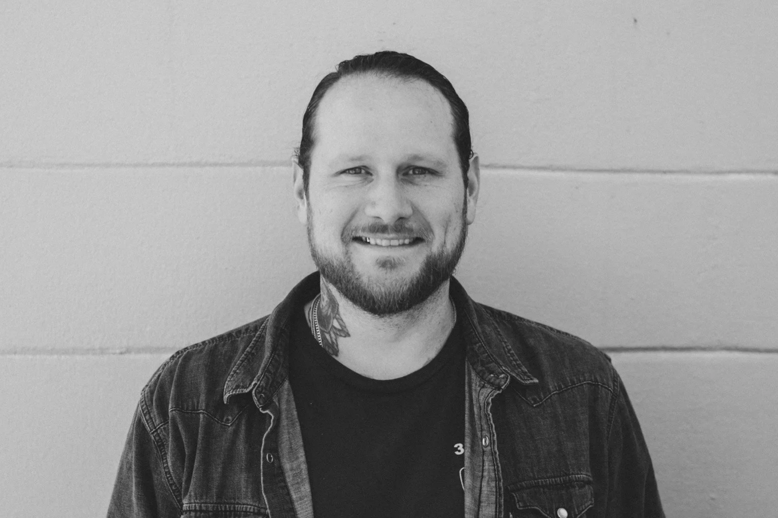 Black and white photo of a smiling man with a beard and tattoos, wearing a denim jacket, standing against a plain wall.