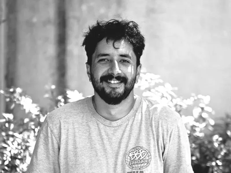 Black and white photo of a cheerful man with dark, curly hair and a beard, wearing a light-colored T-shirt, sitting outdoors with blurred foliage in the background.