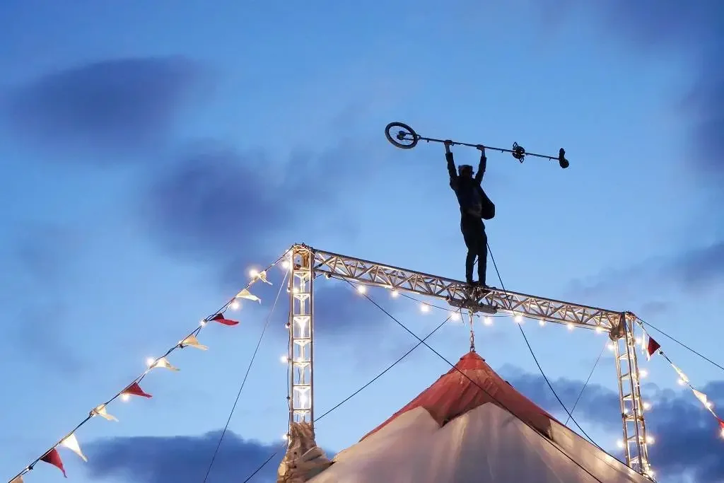 A silhouette of a performer standing on a high wire at a circus, holding a large weather vane with a wheel and arrow, against a evening sky with clouds in Great Yarmouth