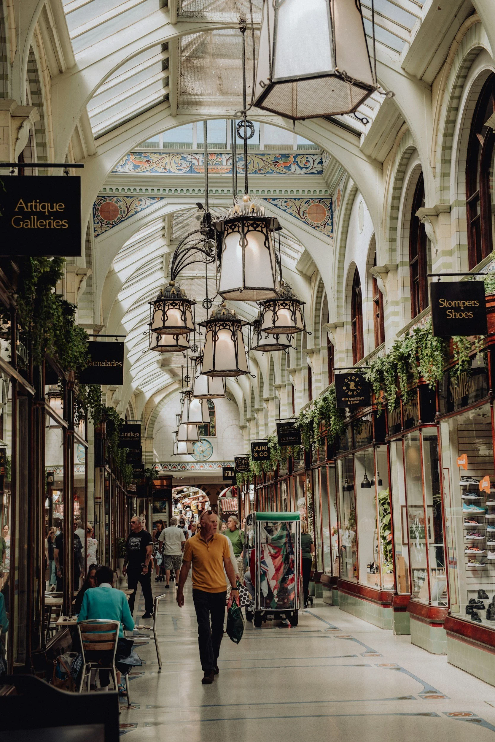 Interior view of a shopping arcade with glass-panelled ceiling, hanging lantern-style lights, and shops on both sides in the Norwich Lanes