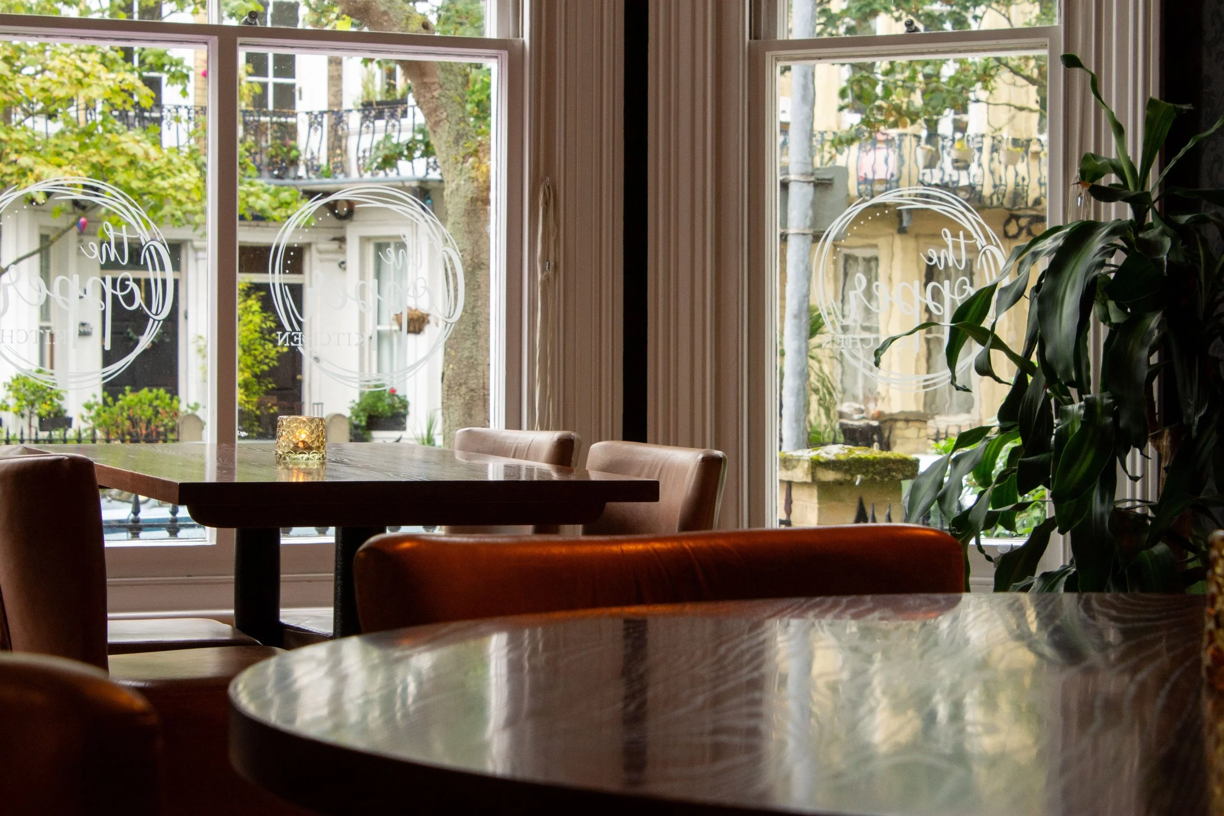 View of the interior of a cozy bar with wooden tables and leather chairs, large windows with a view of a street with buildings, trees, and balconies, and a lit candle on the table.