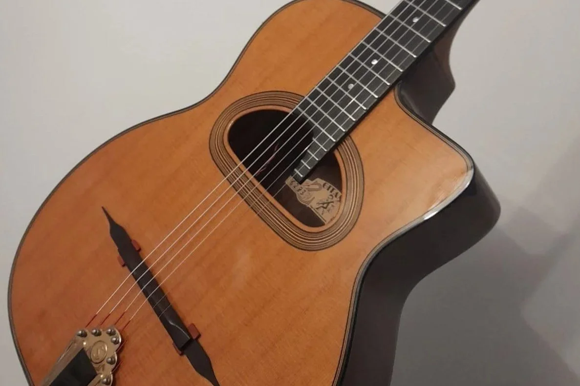 An acoustic guitar with a natural wood finish, six strings, and a sound hole with a label inside. The guitar is resting against a plain wall.