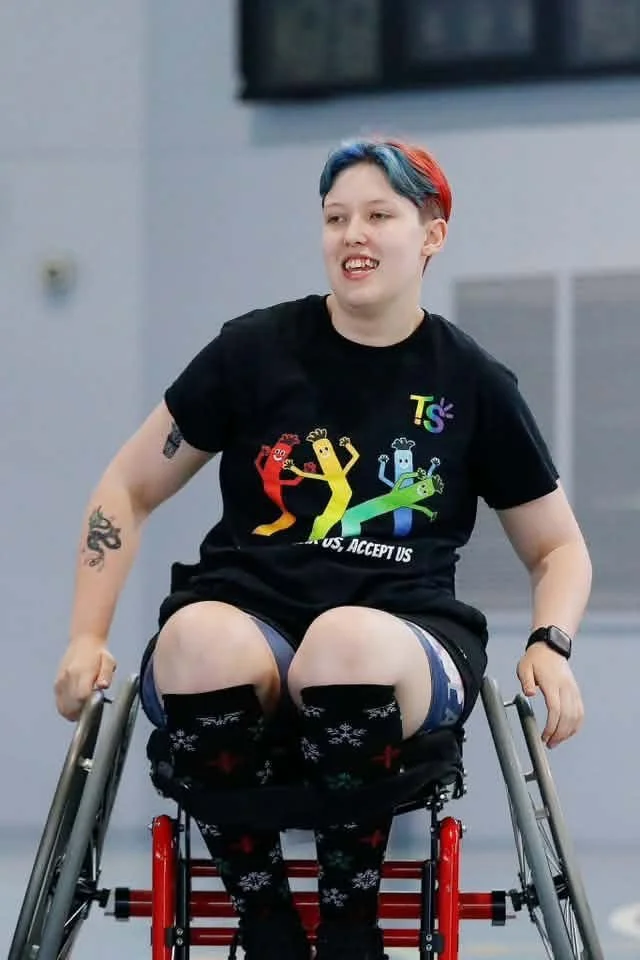 Young person in a wheelchair with colorful hair, wearing a black T-shirt with rainbow colored dancing characters, and festive socks with snowflakes and reindeer, smiling indoors.