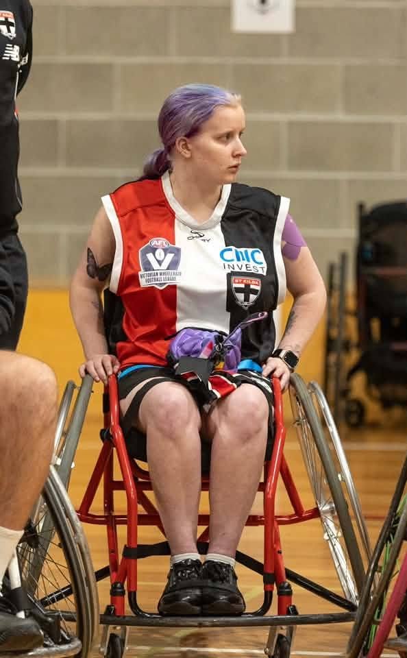 A woman with purple hair sitting in a wheelchair on a court, wearing a sports jersey with various logos, looking to the side.