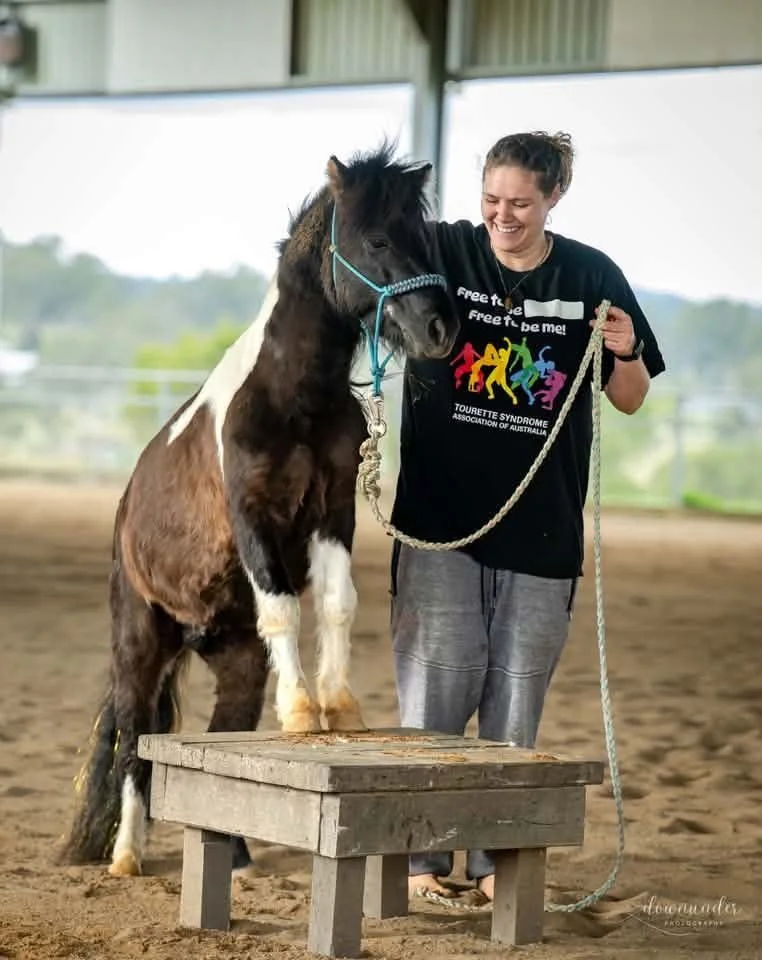 A woman standing next to a small horse with a white and black coat inside a covered riding arena. The woman is smiling and holding a lead rope, while the horse's front legs are on a wooden platform.