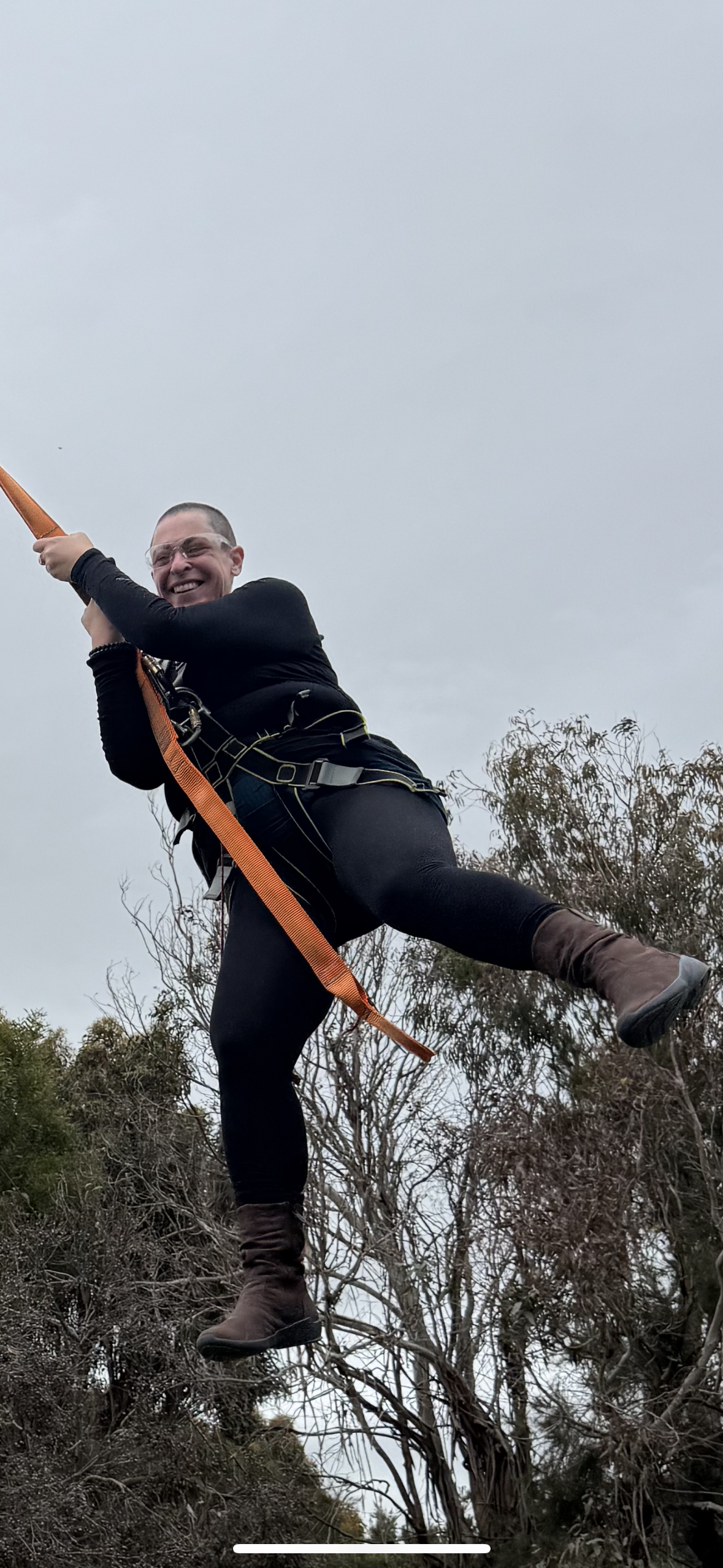 Person wearing glasses and dressed in black climbing a tree with safety harness and orange strap on an overcast day.