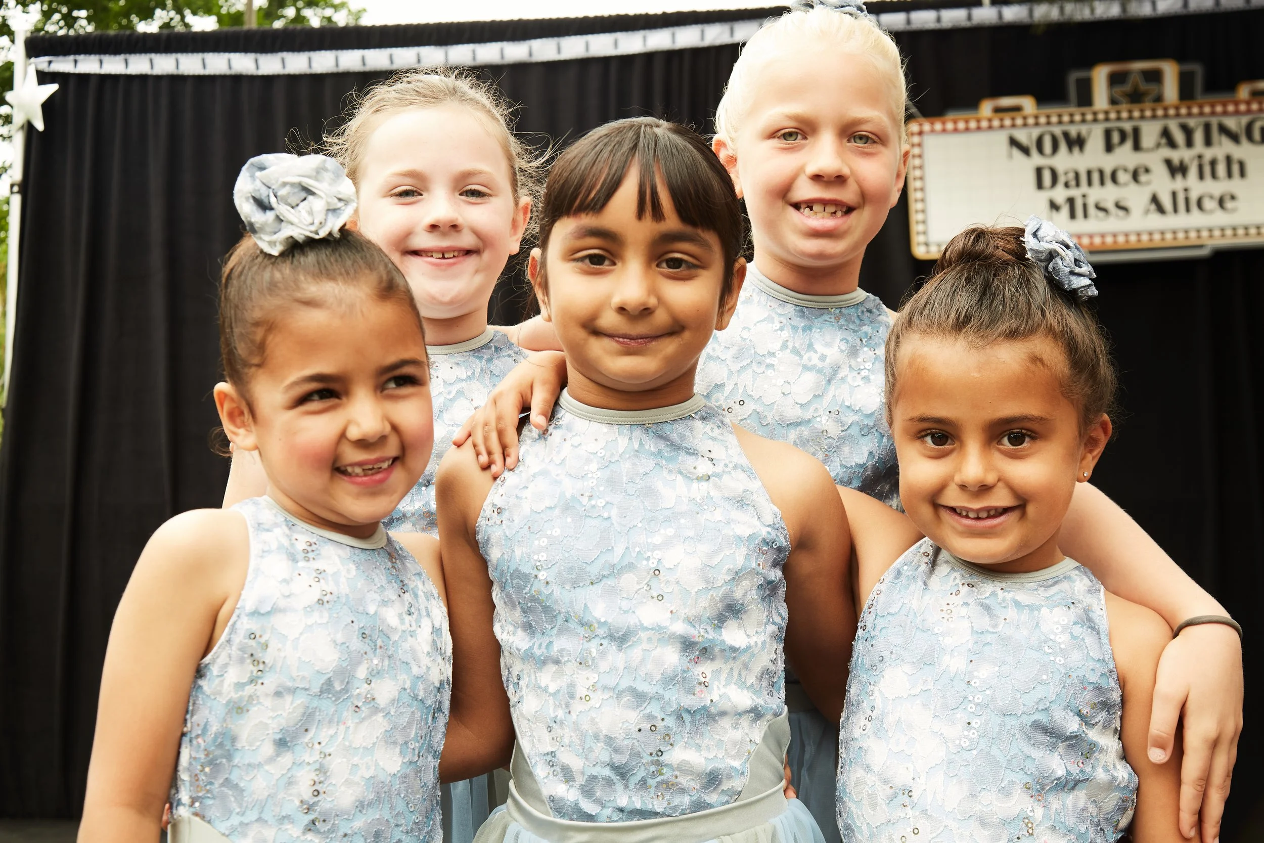 Group of six young girls dressed in light blue dance costumes, posing for a photo in front of a stage with a sign that reads 'Now Playing Dance With Miss Alice'.