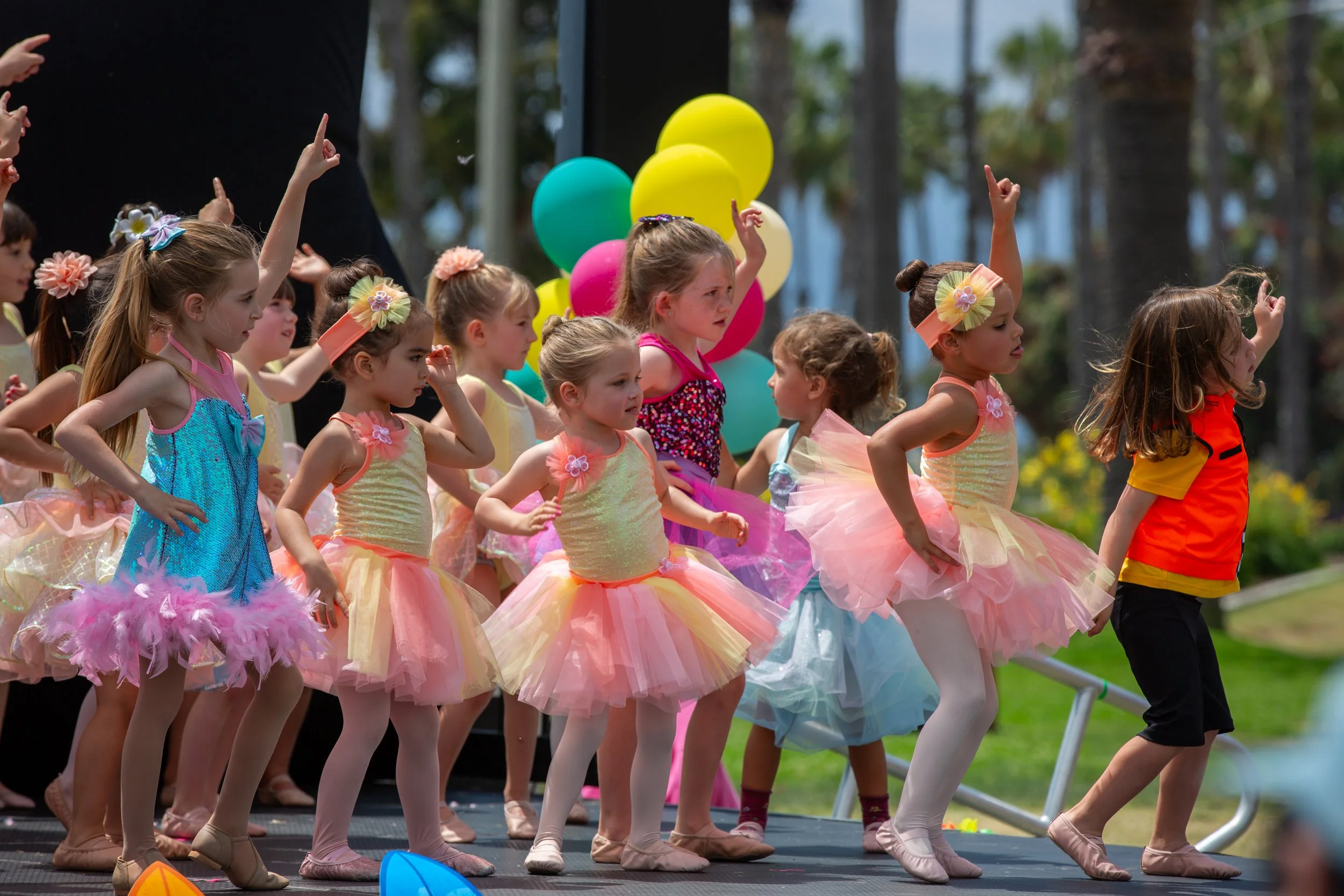 Young children dressed in colorful tutus and ballet costumes perform on an outdoor stage, with balloons in the background.