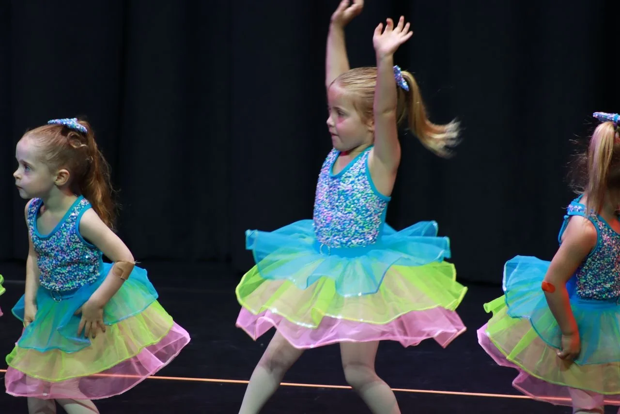 Three young girls in colorful tutu dresses performing a dance on stage with black background.