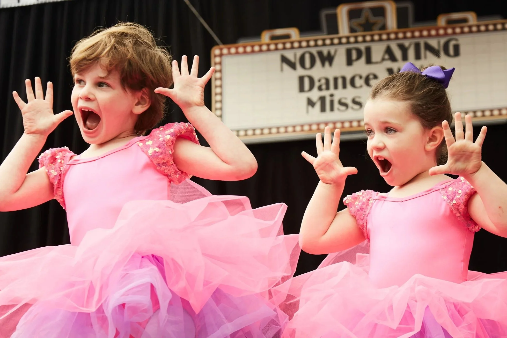 Two young girls dressed as ballerinas with pink tutus and matching pink tops, singing or performing on stage with a marquee sign in the background that reads 'Now Playing Dance Miss'.