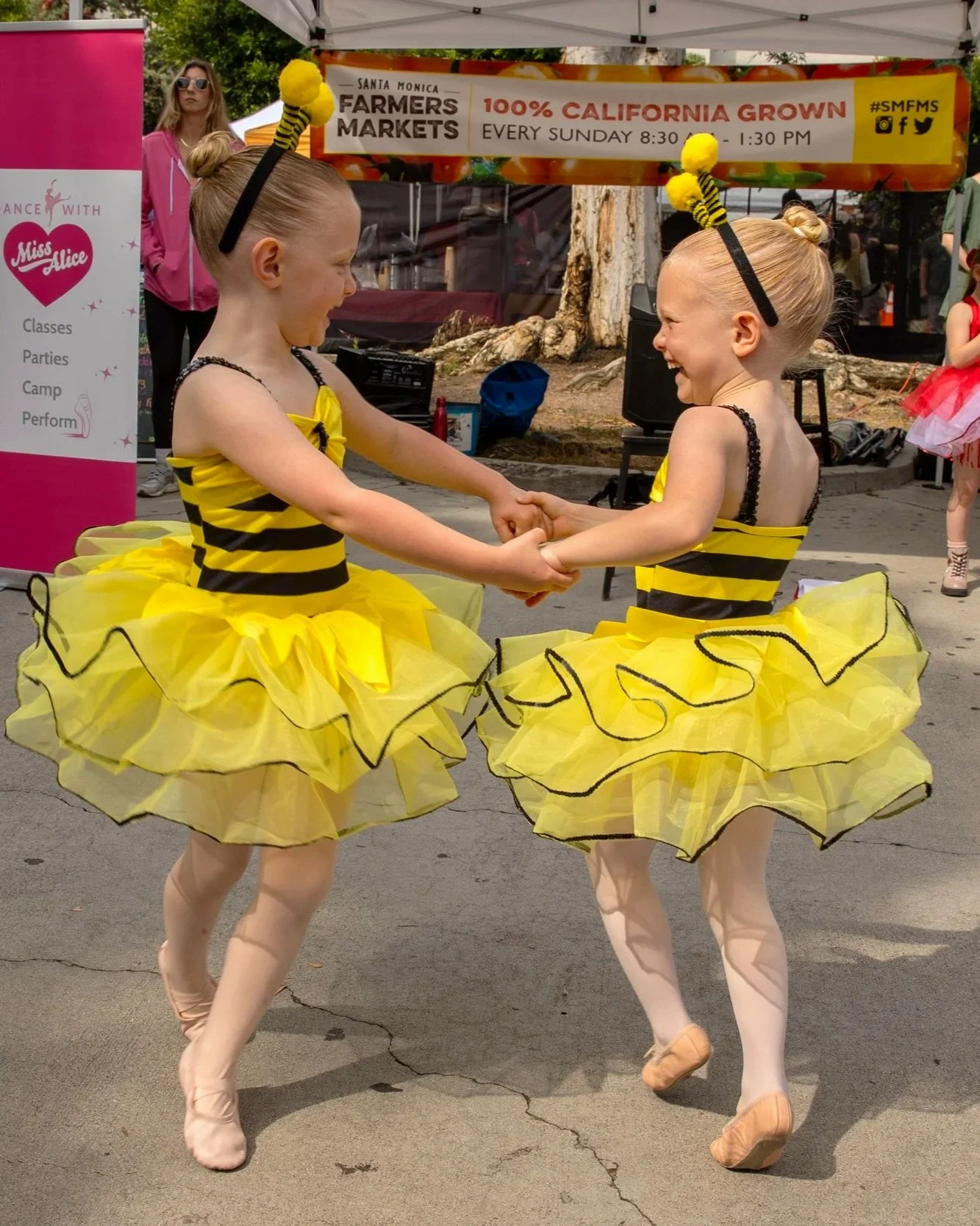 Two young girls dressed as bees in yellow and black striped costumes with tutu skirts, laughing and holding hands while dancing outdoors.
