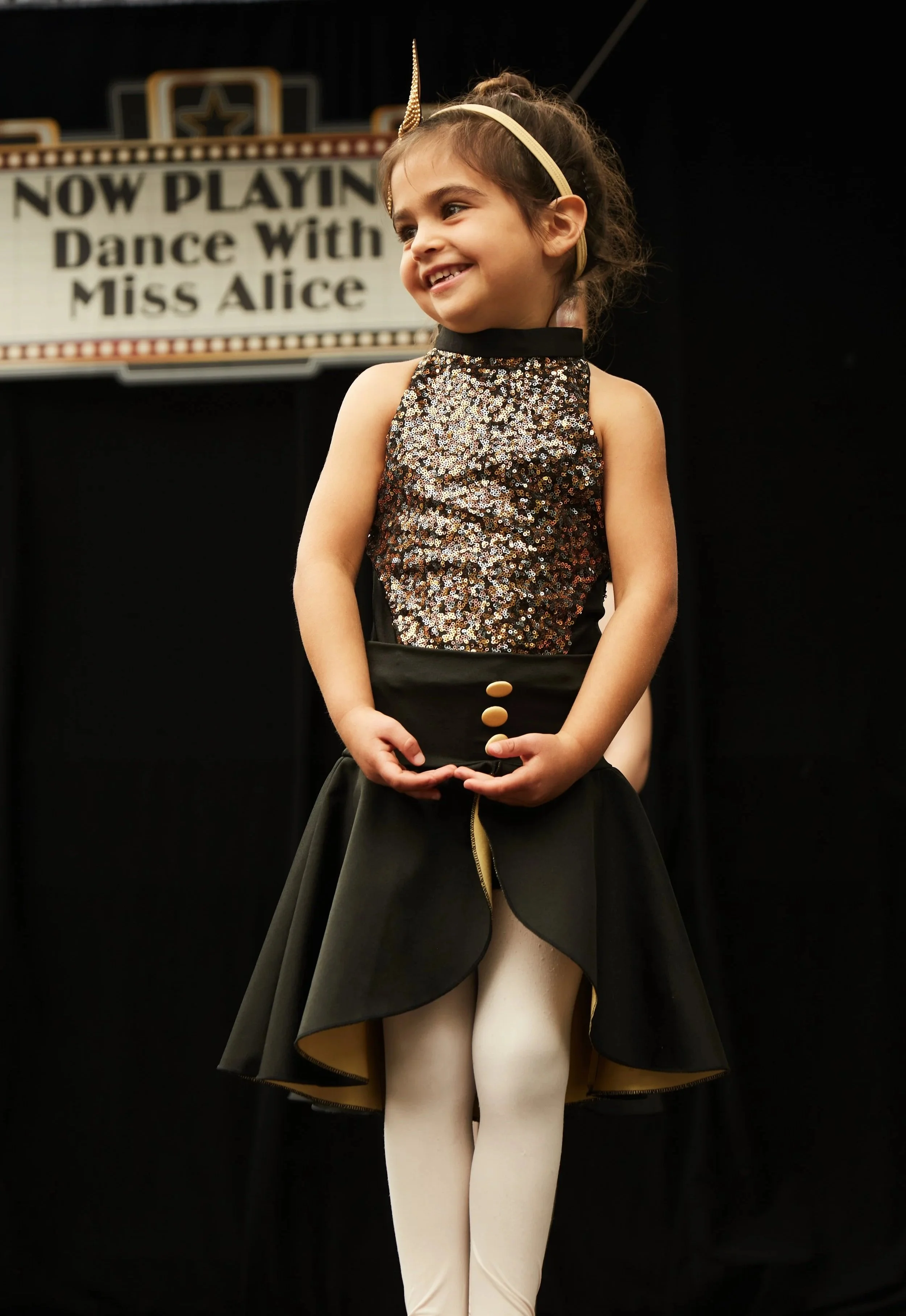 Young girl in a sparkly top and black skirt, wearing a unicorn headband, standing on stage with a sign behind her that says 'Now Playing: Dance With Miss Alice'.
