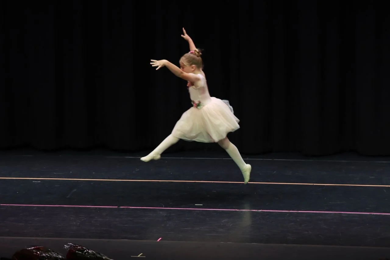 Young girl in a ballet tutu performing a dance leap on stage with black curtains in the background.