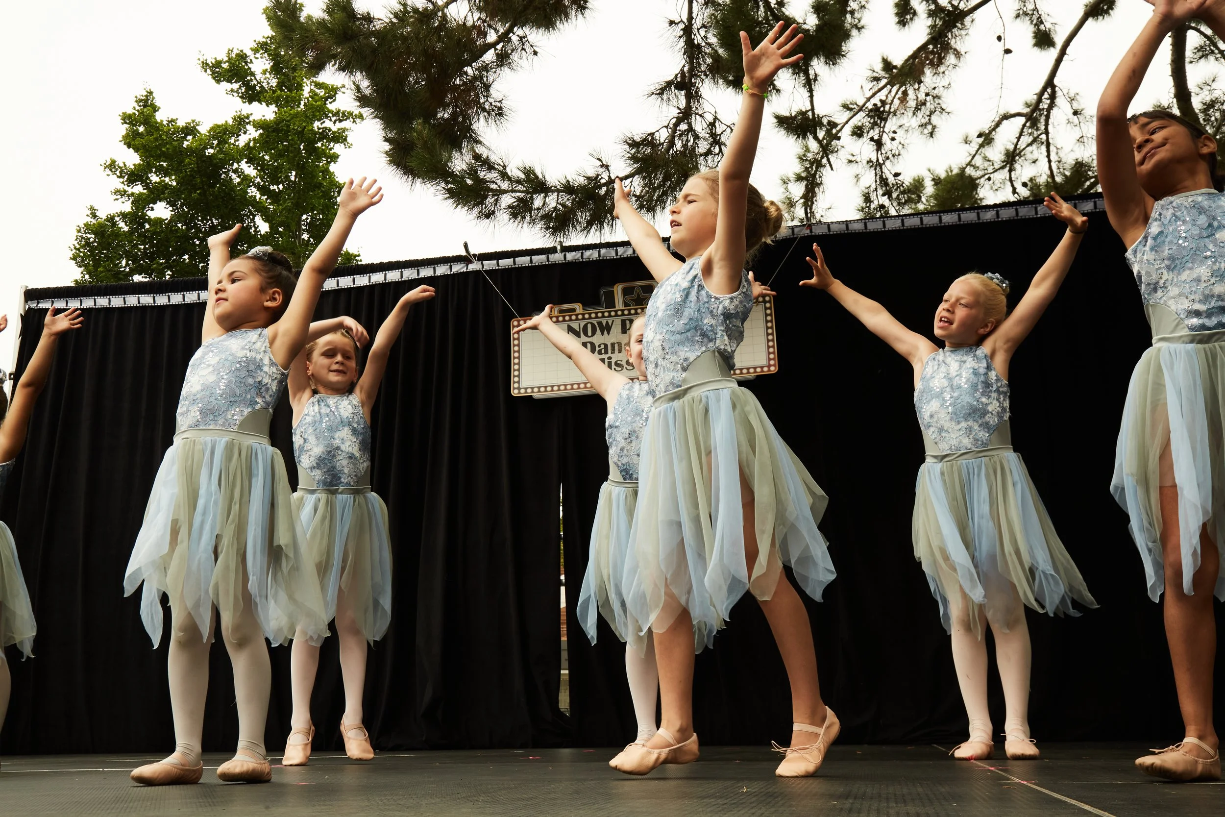 Young girls dressed in blue ballet costumes performing a dance on stage outdoors with black curtains and trees in the background.