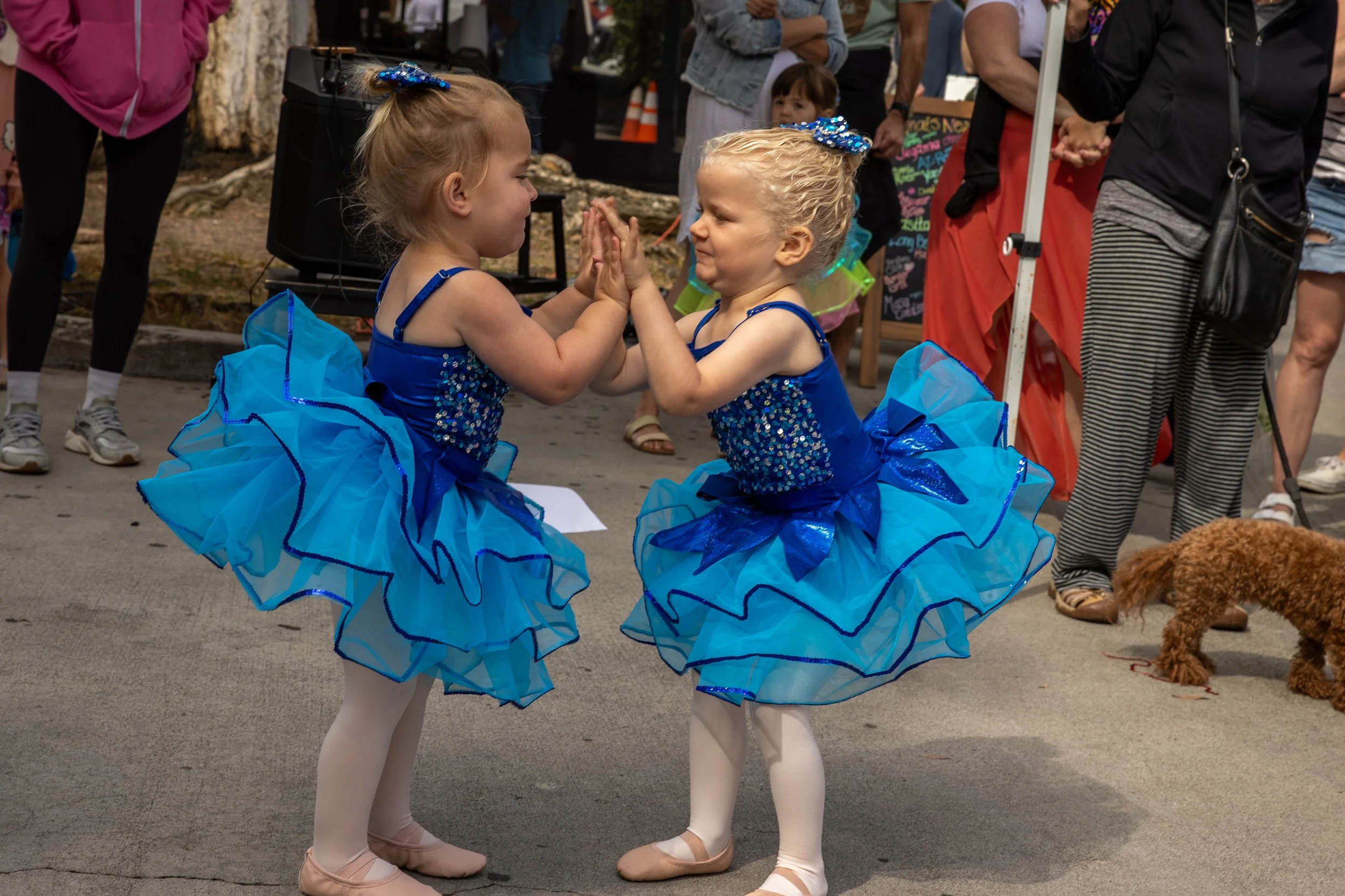 Two young girls in blue tutus and ballet shoes are playing a hand game on a sidewalk, with onlookers and a small dog in the background.