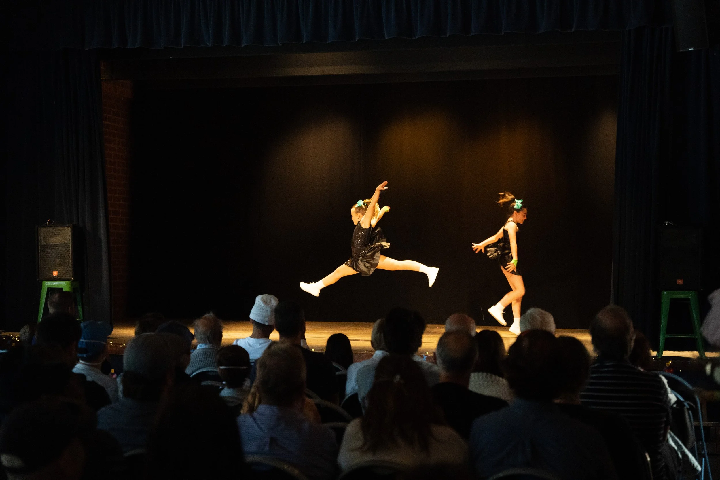 Two young girls are performing a tap dance on stage, dressed in black with turquoise bows, white socks, and tap shoes, catching the audience's attention.