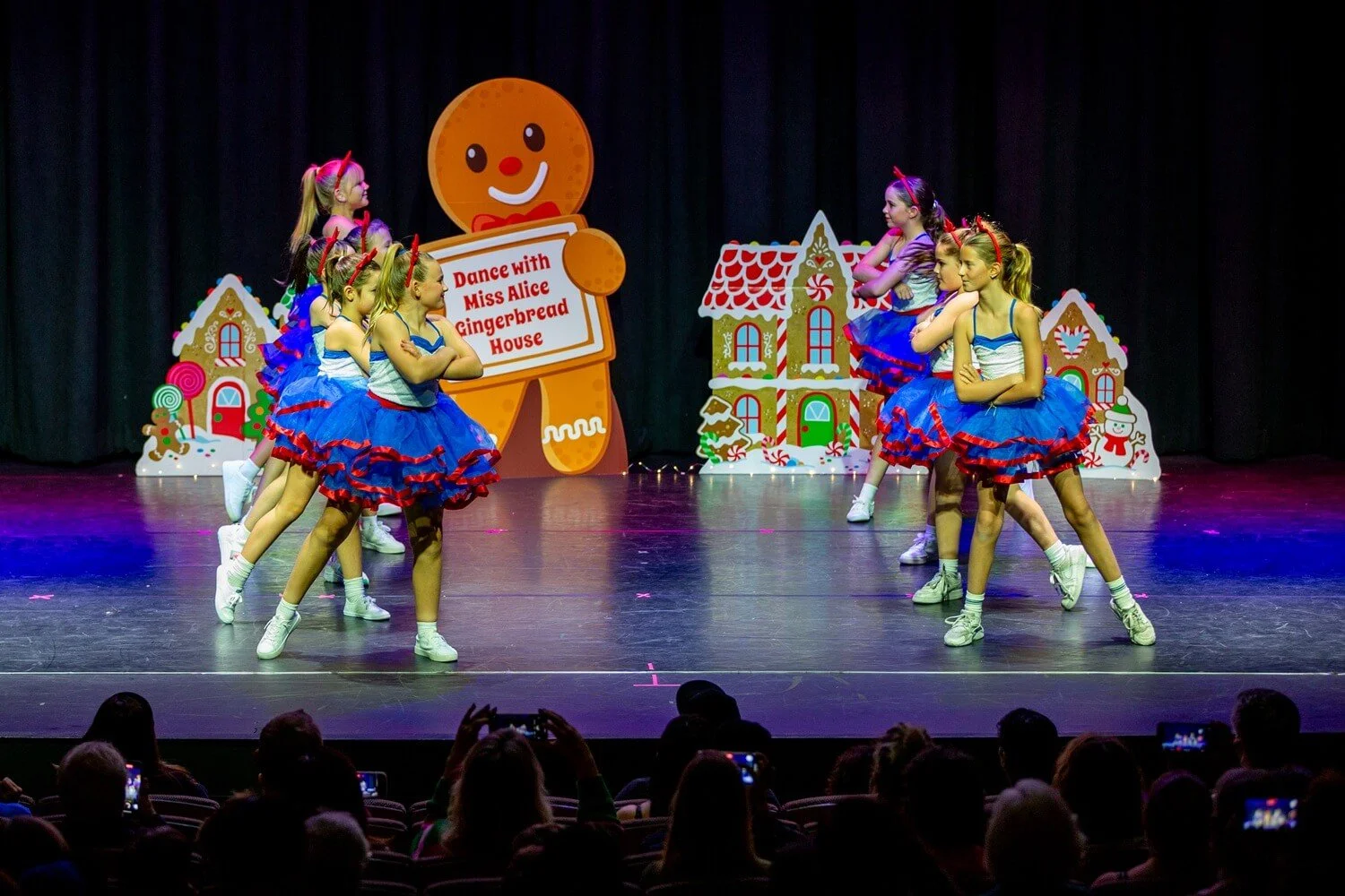 Young girls in colorful costumes perform a dance on stage decorated with gingerbread house and holiday-themed props during a Christmas show.