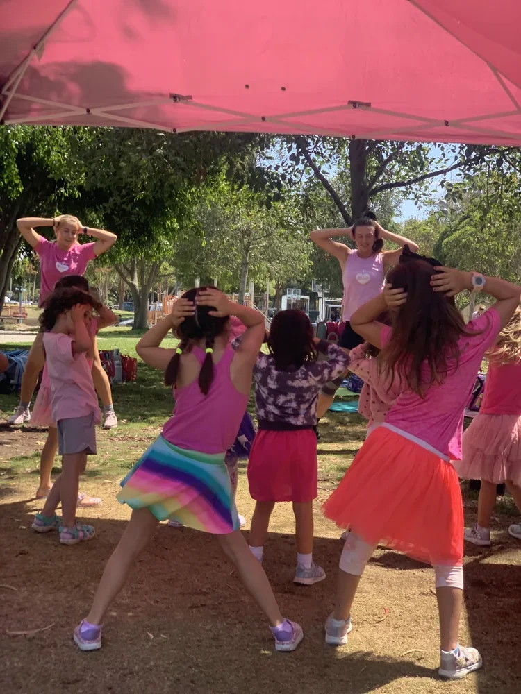 Children under a pink tent participating in a dance or exercise activity in a park with trees, some with hands on their heads, wearing colorful casual clothes.