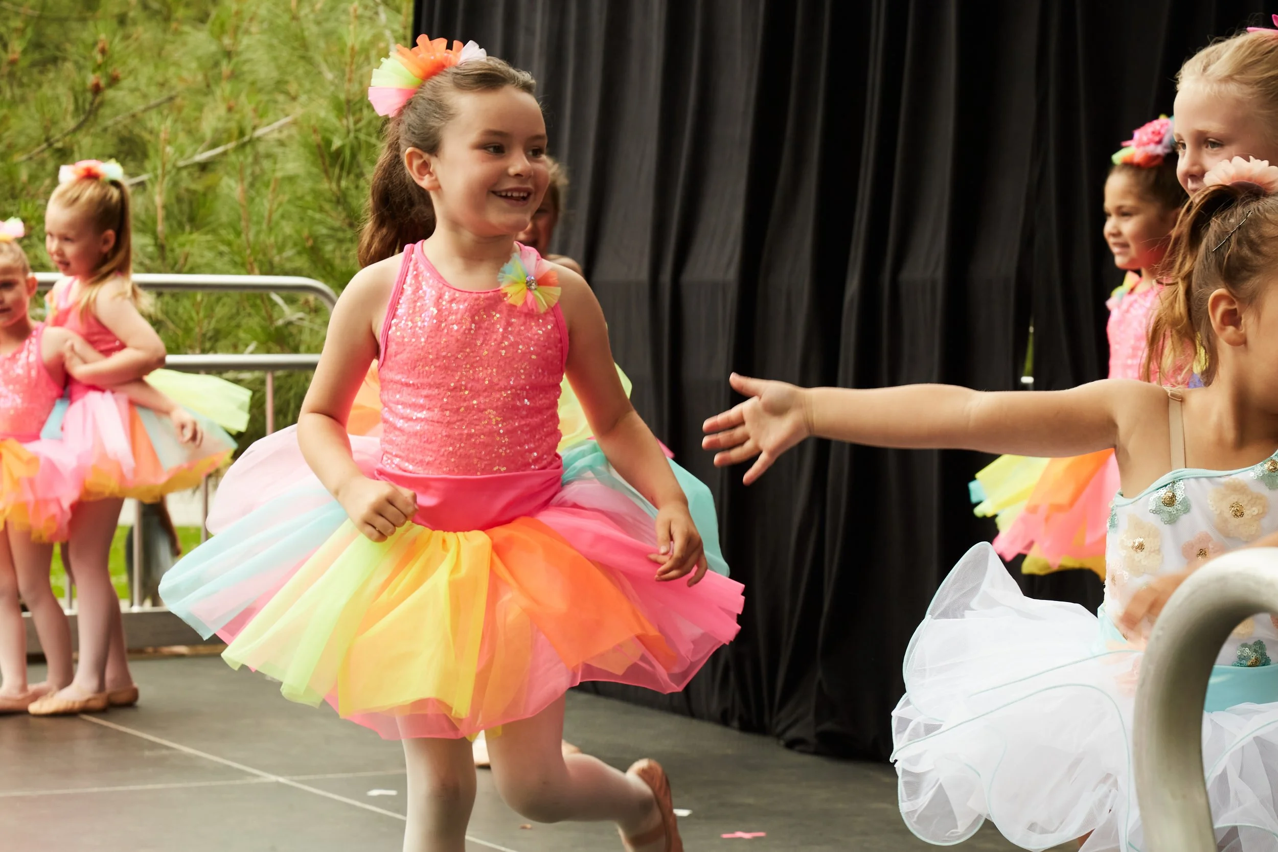 Children dressed in colorful tutus and ballet costumes participating in a dance recital or rehearsal on a stage with a black curtain backdrop.