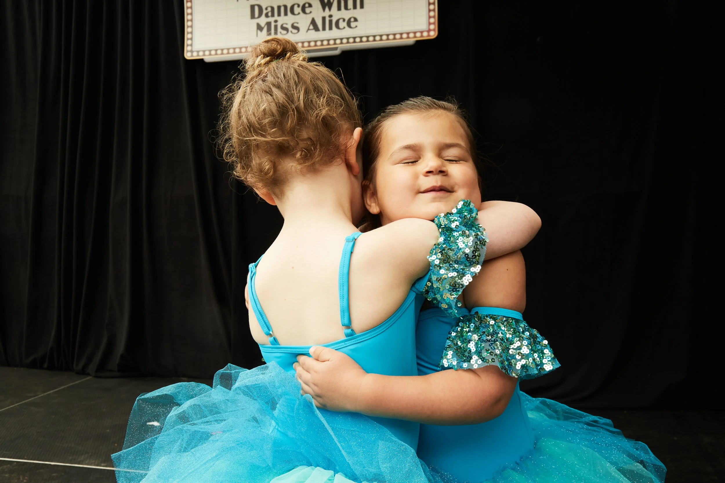 Two young girls in blue dance costumes hugging each other on stage with a sign in the background that reads 'Dance With Miss Alice'.