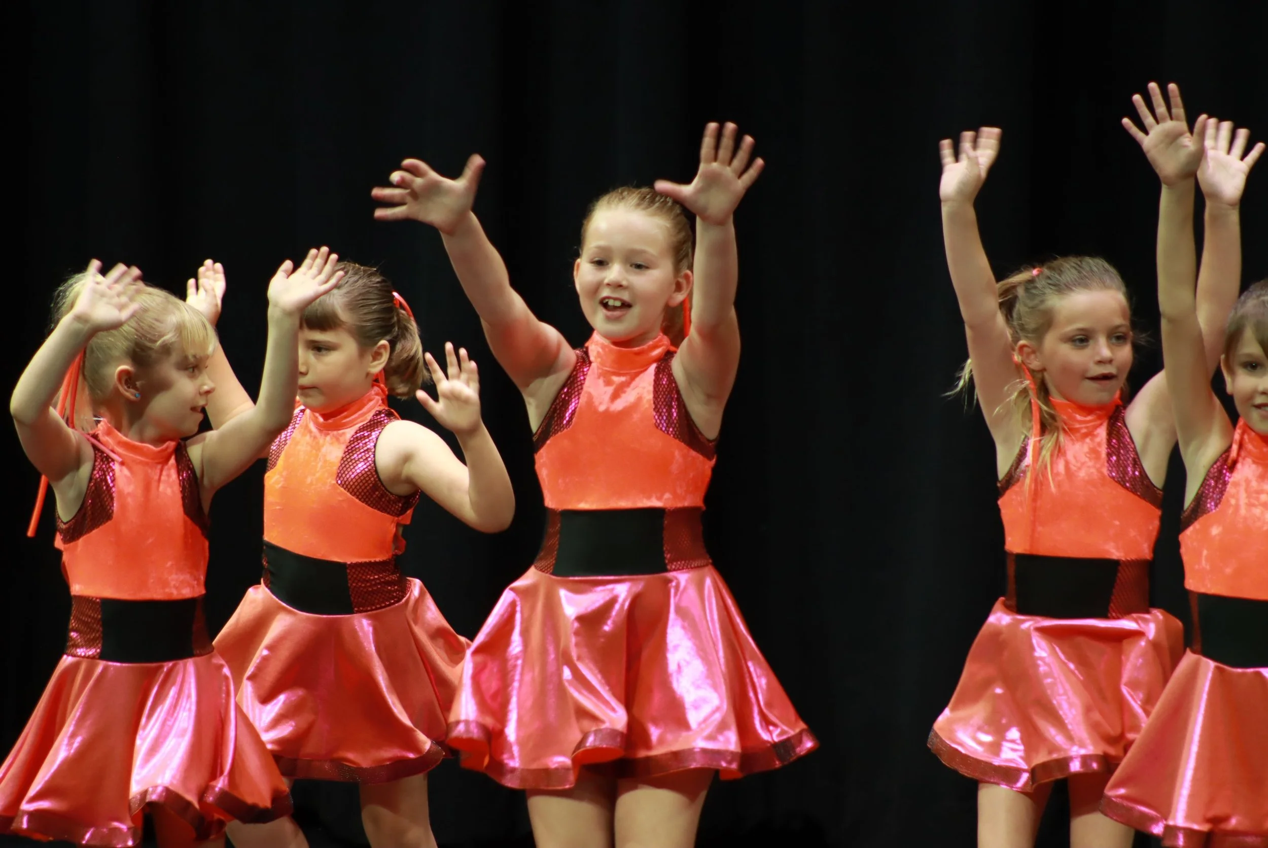 Young girls performing a dance on stage, wearing matching orange and pink costumes with black accents, raising their hands during a performance.