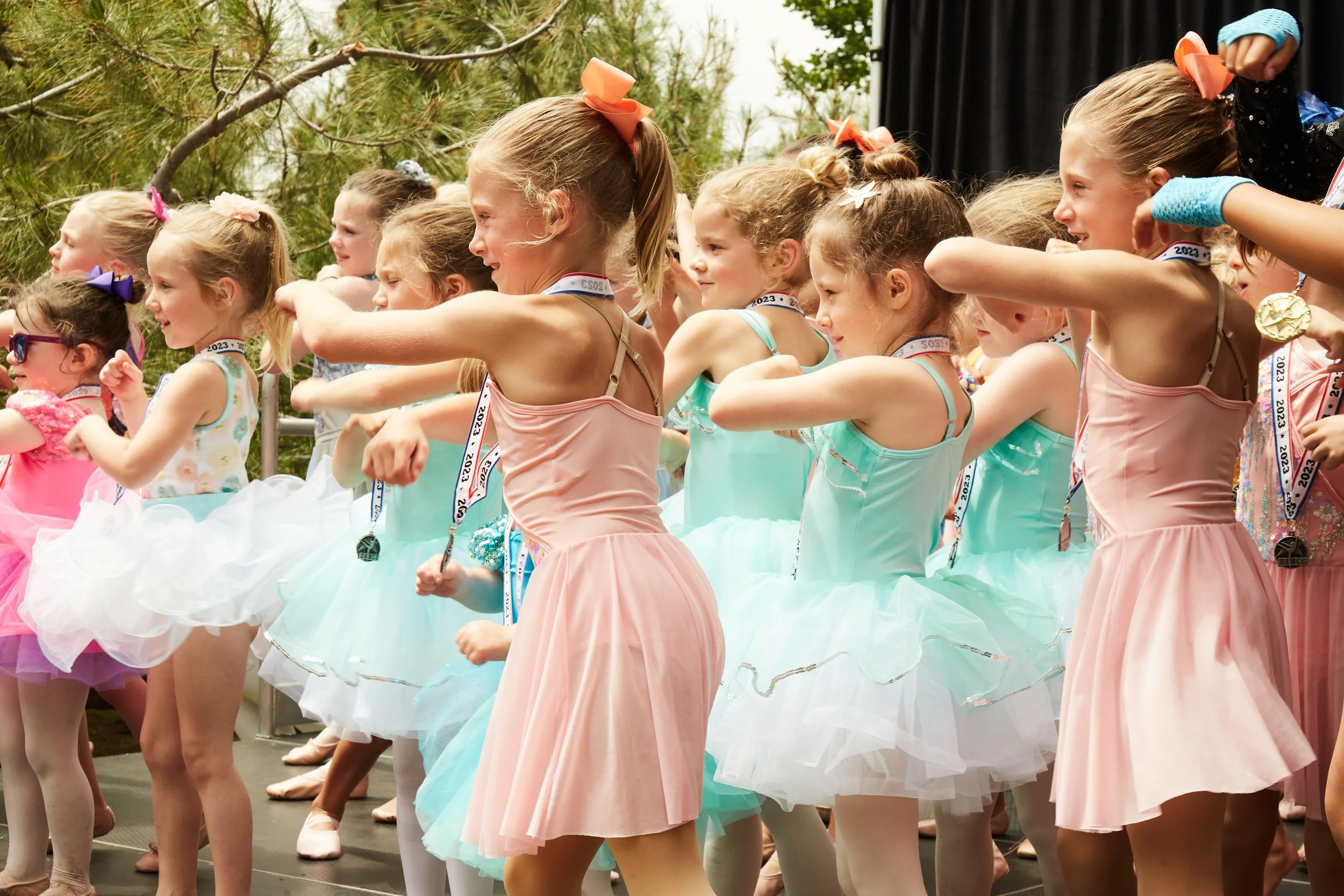 Group of young girls dressed in colorful ballet costumes performing on stage outdoors.