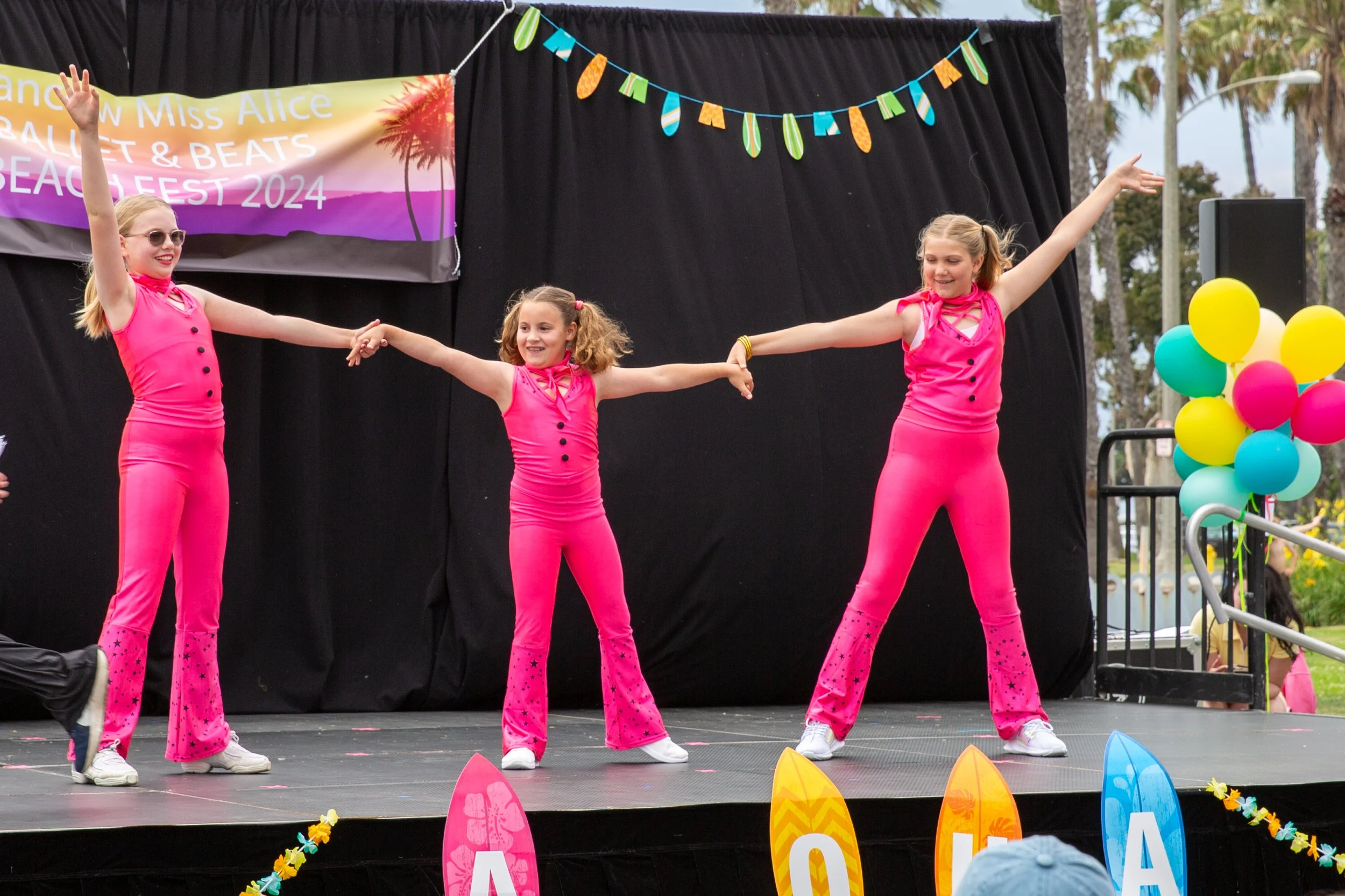 Three young girls in bright pink dance costumes performing on a stage at an outdoor beach festival, with colorful balloons and banners in the background.